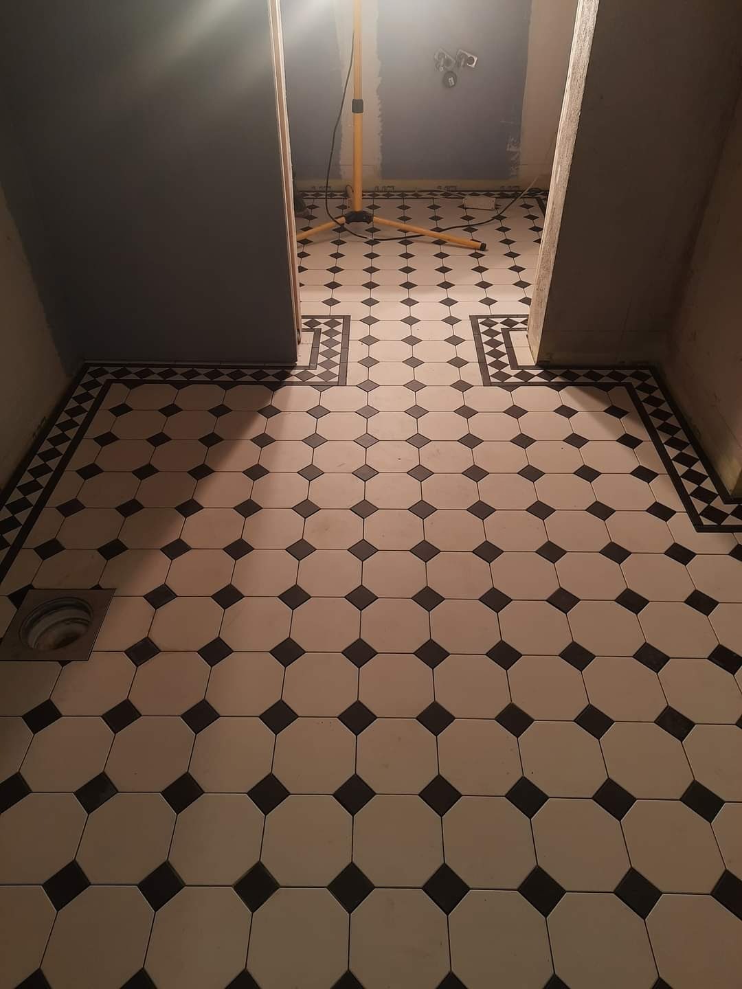 Ułożona podłoga z płytek w stylu retro, biało-czarne oktagonalne płytki z czarnymi kwadratami, obramowanie z dekoracyjnej mozaiki, widoczny odpływ podłogowy, w tle lampa robocza na trójnogu...