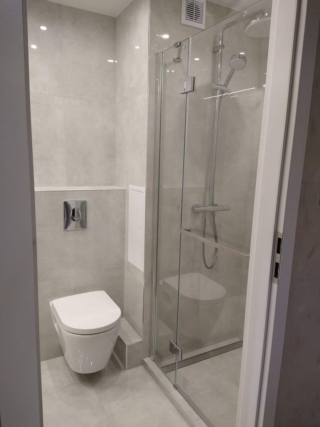 Nowoczesna łazienka z kabiną prysznicową typu walk-in, podwieszaną toaletą i szarymi płytkami na ścianach i podłodze, widoczny fragment wentylacji na suficie.