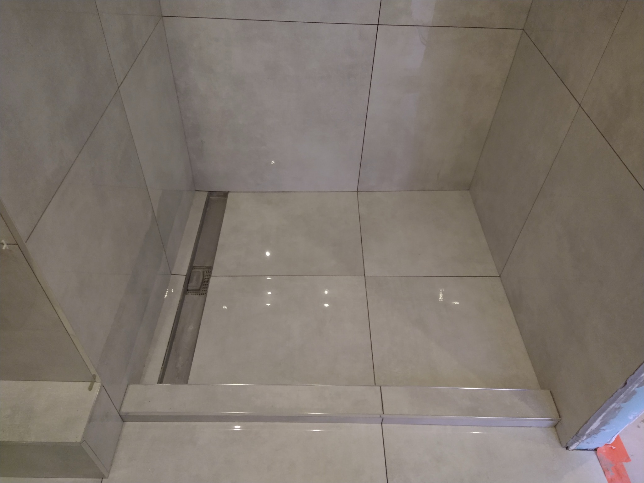 Wykończony brodzik prysznicowy z odpływem liniowym, wyłożony jasnymi, kwadratowymi płytkami ceramicznymi z delikatnym wzorem, widoczne fugi i listwy wykończeniowe.