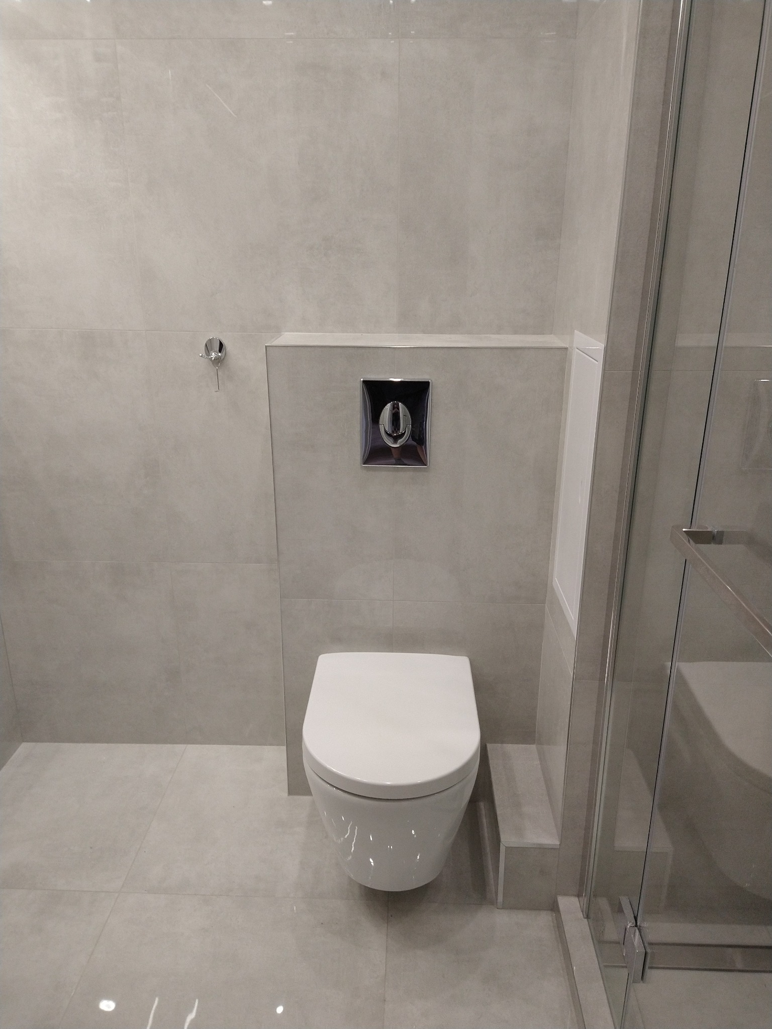 Nowoczesna łazienka z wiszącą toaletą, szarą glazurą i kabiną prysznicową ze szklanymi drzwiami, widoczny chromowany wieszak na ręcznik i przycisk spłuczki.