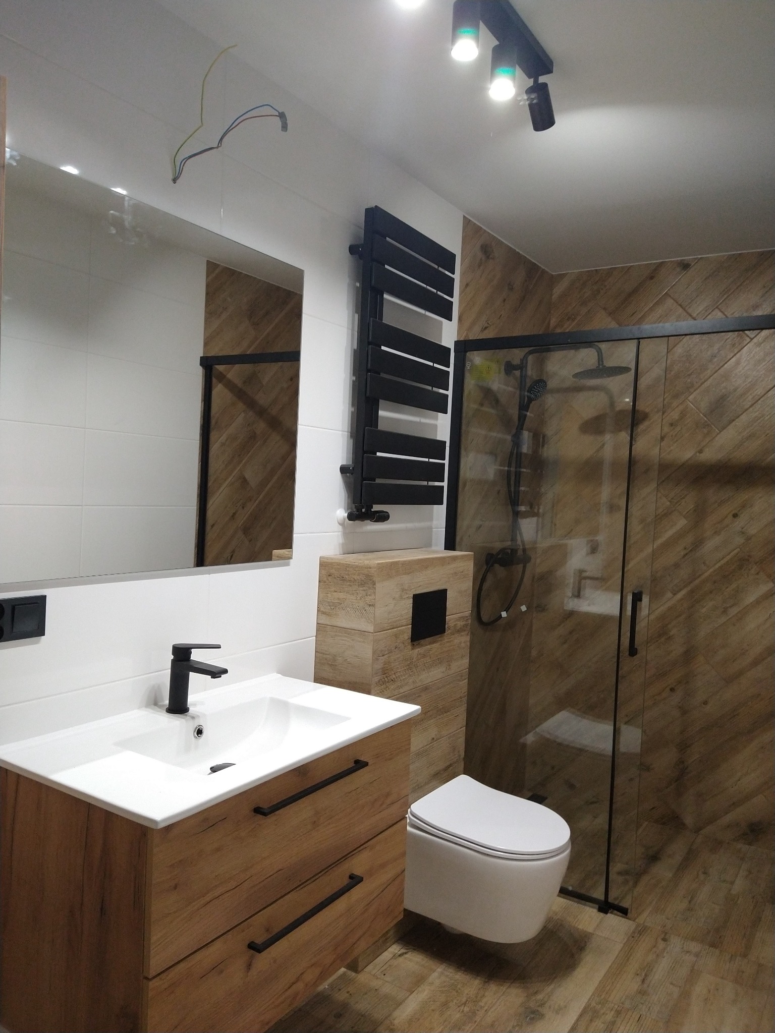 Nowoczesna łazienka z drewnopodobnymi płytkami na ścianach i podłodze, czarna armatura, umywalka z drewnianą szafką, kabina prysznicowa z czarnymi ramami, wisząca toaleta i czarny grzejnik...