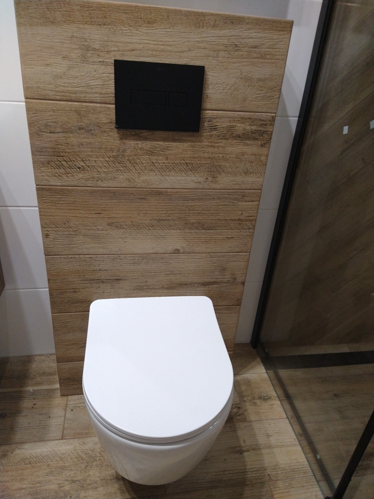 Nowoczesna toaleta podwieszana z czarnym przyciskiem spłukującym na ścianie wyłożonej płytkami imitującymi drewno, widoczny fragment kabiny prysznicowej.