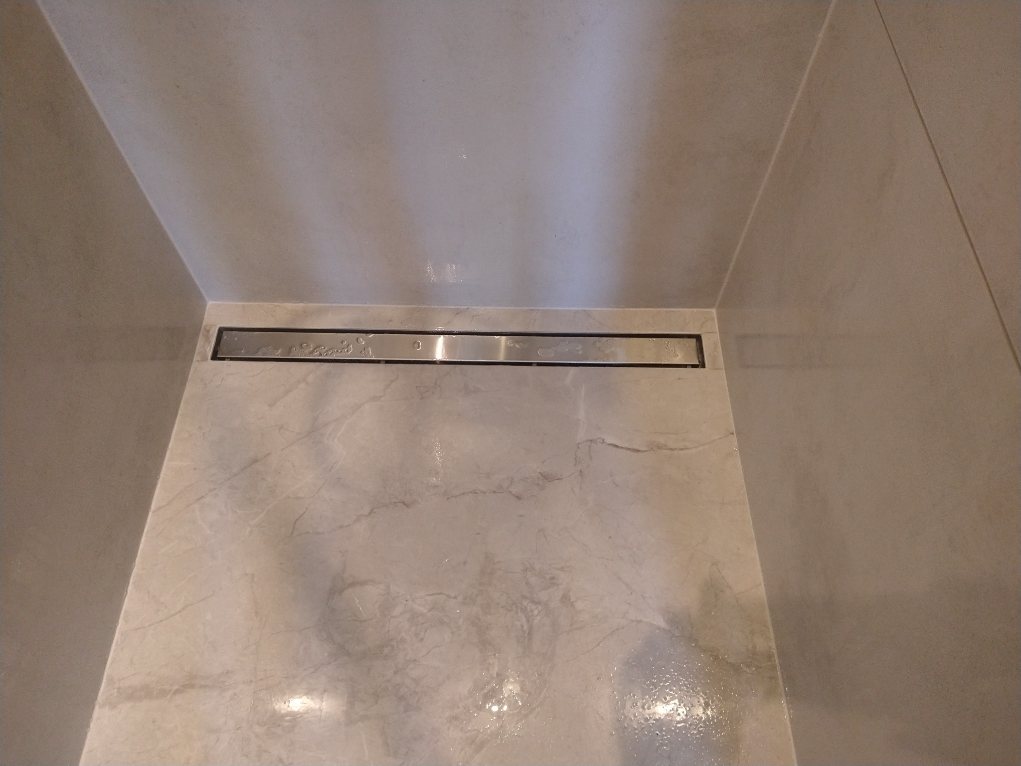 Nowoczesny odpływ liniowy ze stali nierdzewnej w kabinie prysznicowej wyłożonej płytkami imitującymi marmur, widoczne drobne krople wody na powierzchni płytek.