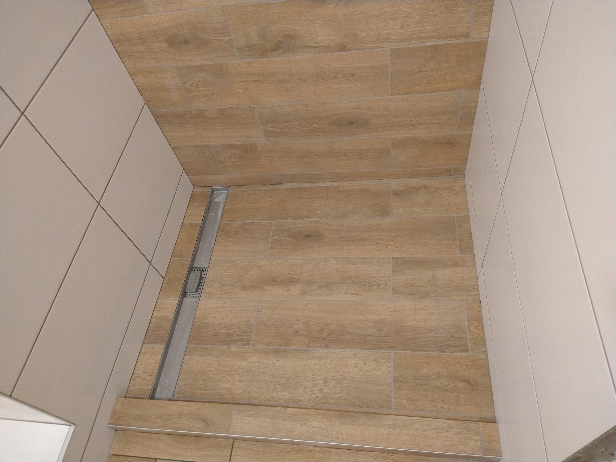 Nowoczesna kabina prysznicowa z odpływem liniowym, wyłożona płytkami imitującymi drewno na podłodze i jasnymi, kwadratowymi płytkami na ścianach.