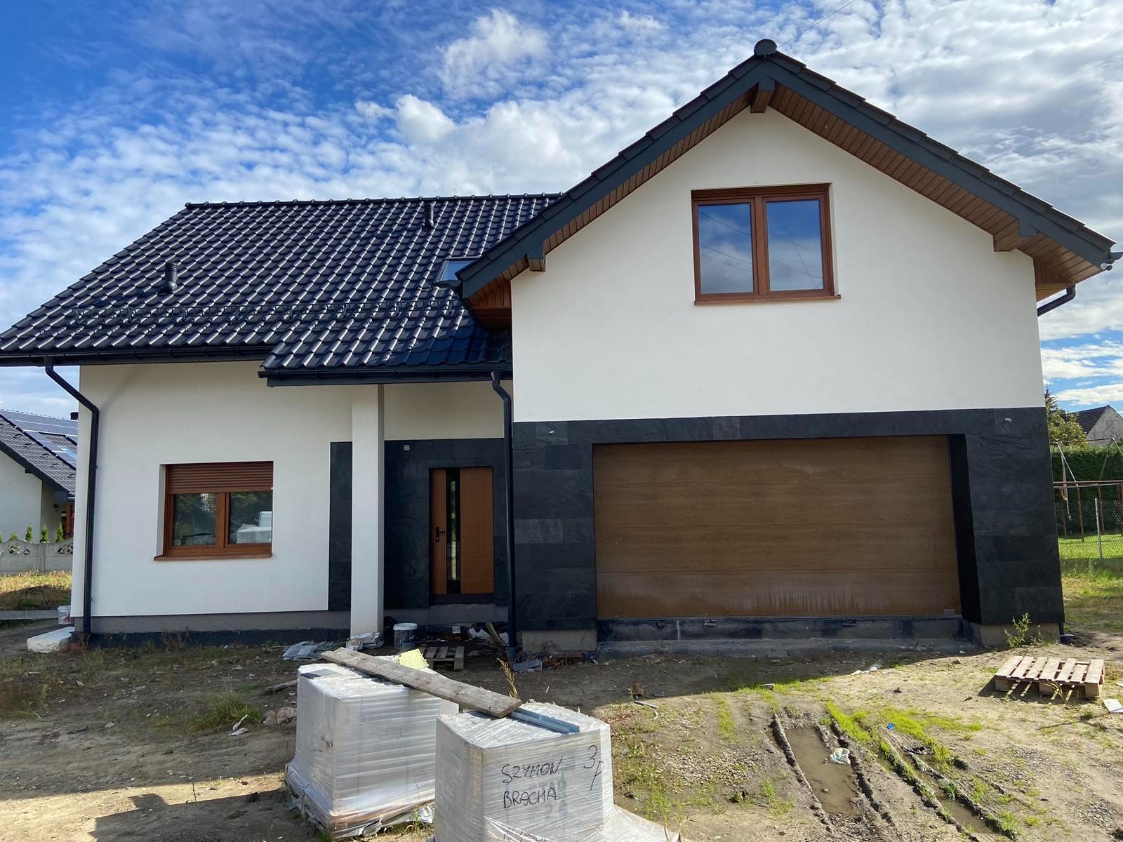 Nowoczesny dom jednorodzinny z białą elewacją, czarnym dachem i elementami z ciemnego kamienia, widoczne pakiety cegieł na podwórku.