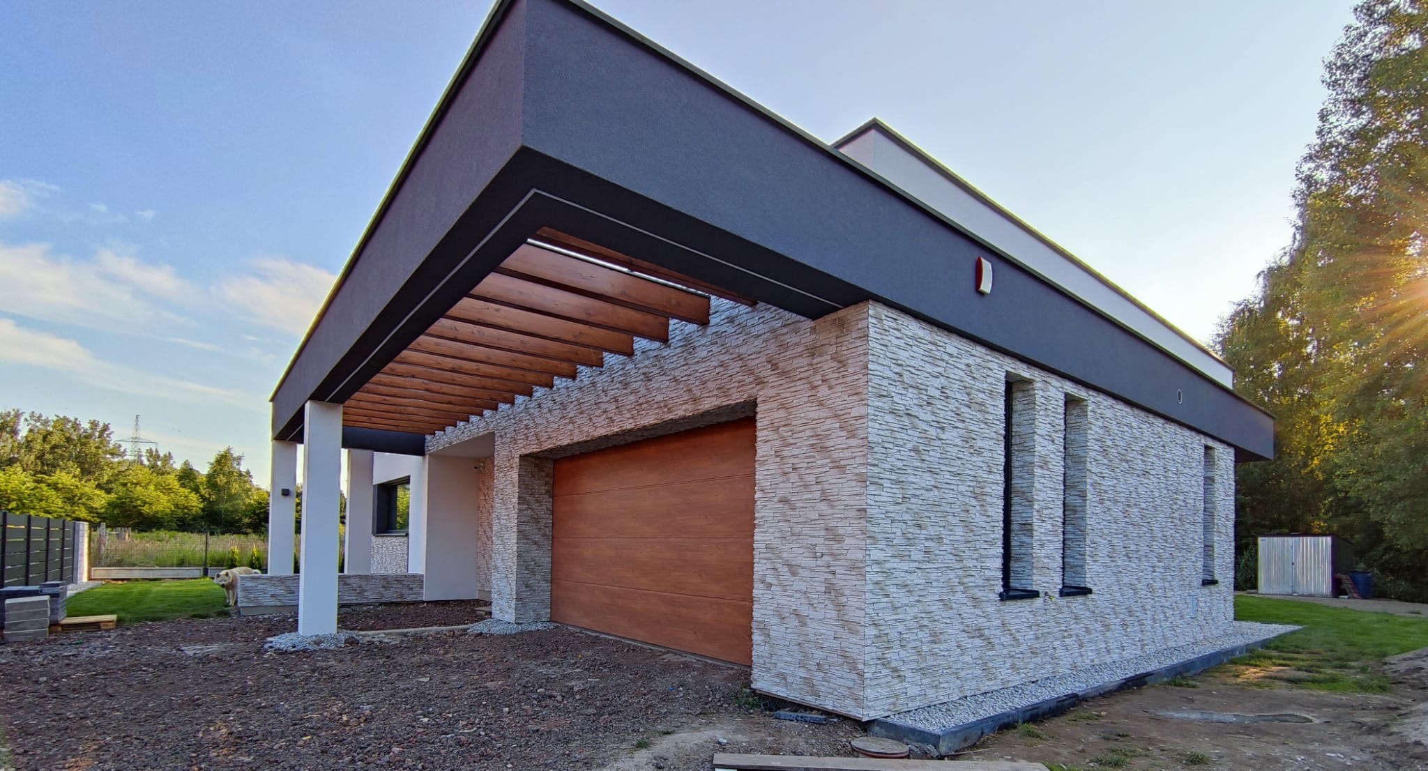 Nowoczesny dom jednorodzinny z elewacją z kamienia dekoracyjnego i drewnianą bramą garażową, widok z zewnątrz w słoneczny dzień.
