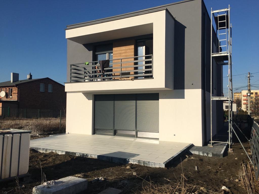 Nowoczesny dom z szarą i białą elewacją, balkonem z metalową balustradą i rusztowaniem przy ścianie, widok z zewnątrz w słoneczny dzień.