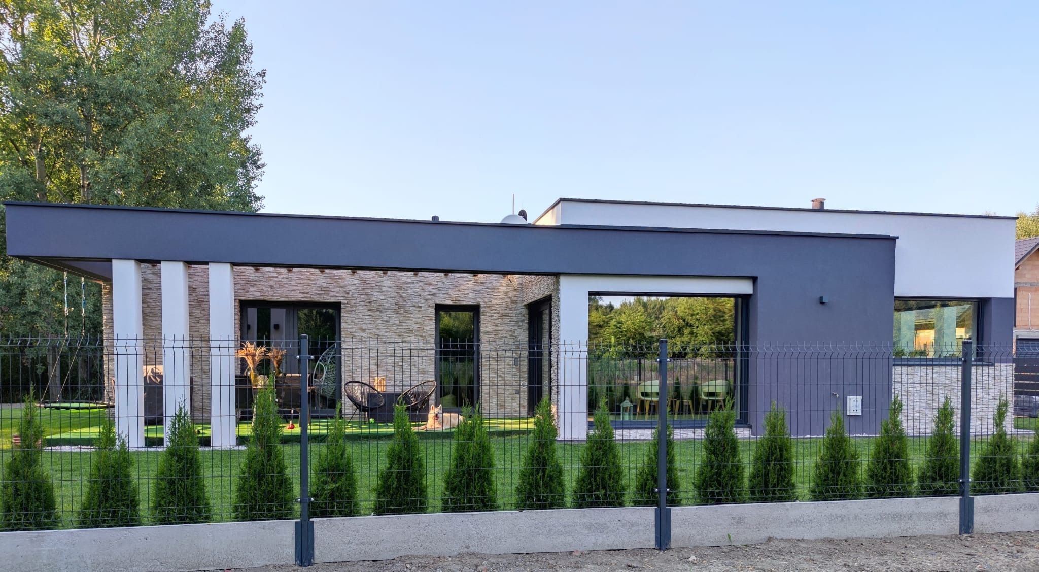 Nowoczesny dom jednorodzinny z elewacją z kamienia dekoracyjnego, ciemnoszarą stolarką okienną i metalowym ogrodzeniem panelowym z niskimi tujami.