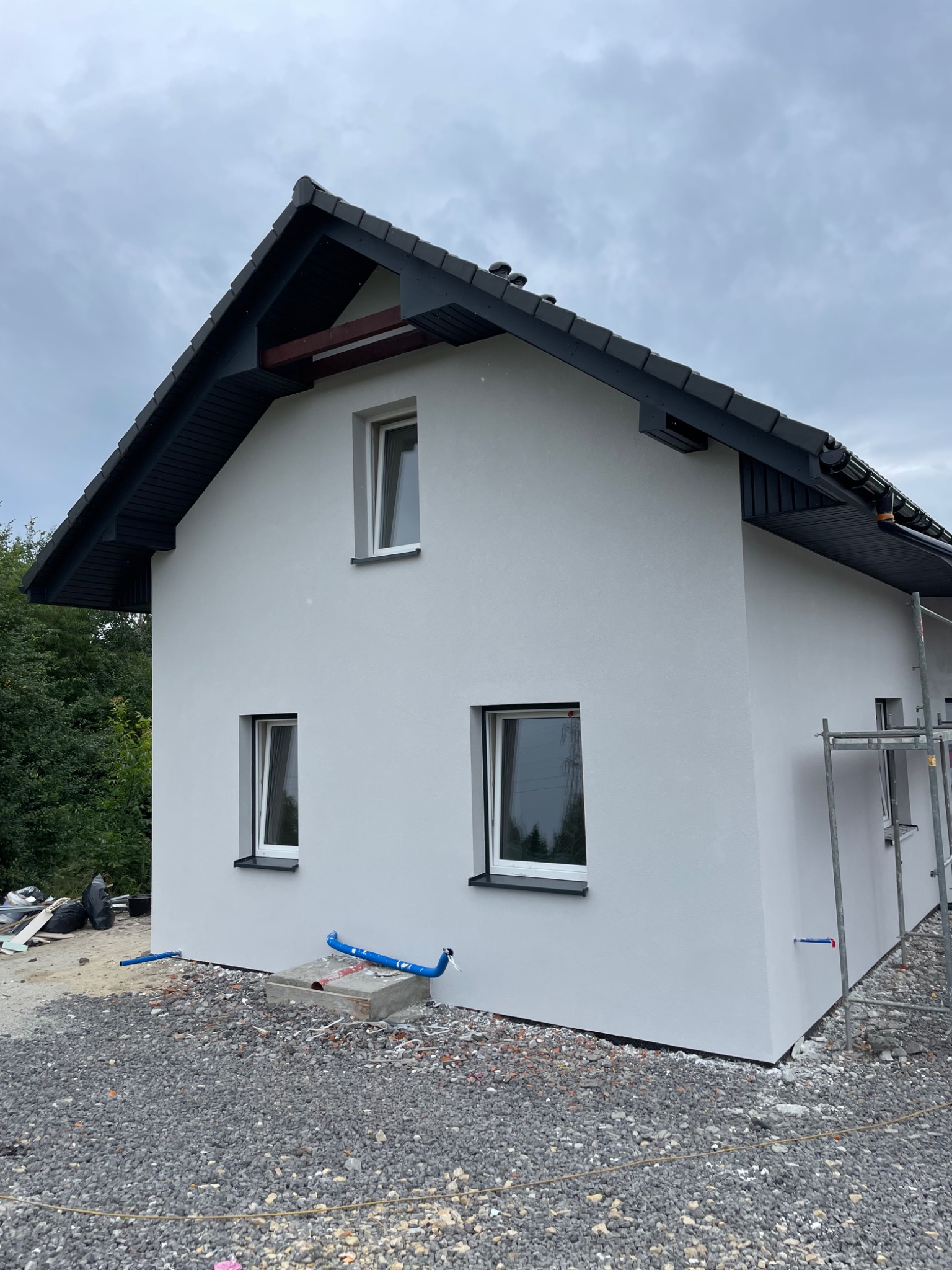 Świeżo otynkowany, biały dom jednorodzinny z ciemną dachówką, widok z boku, na ziemi gruz i niebieskie rury, w tle zieleń drzew, pochmurne niebo.