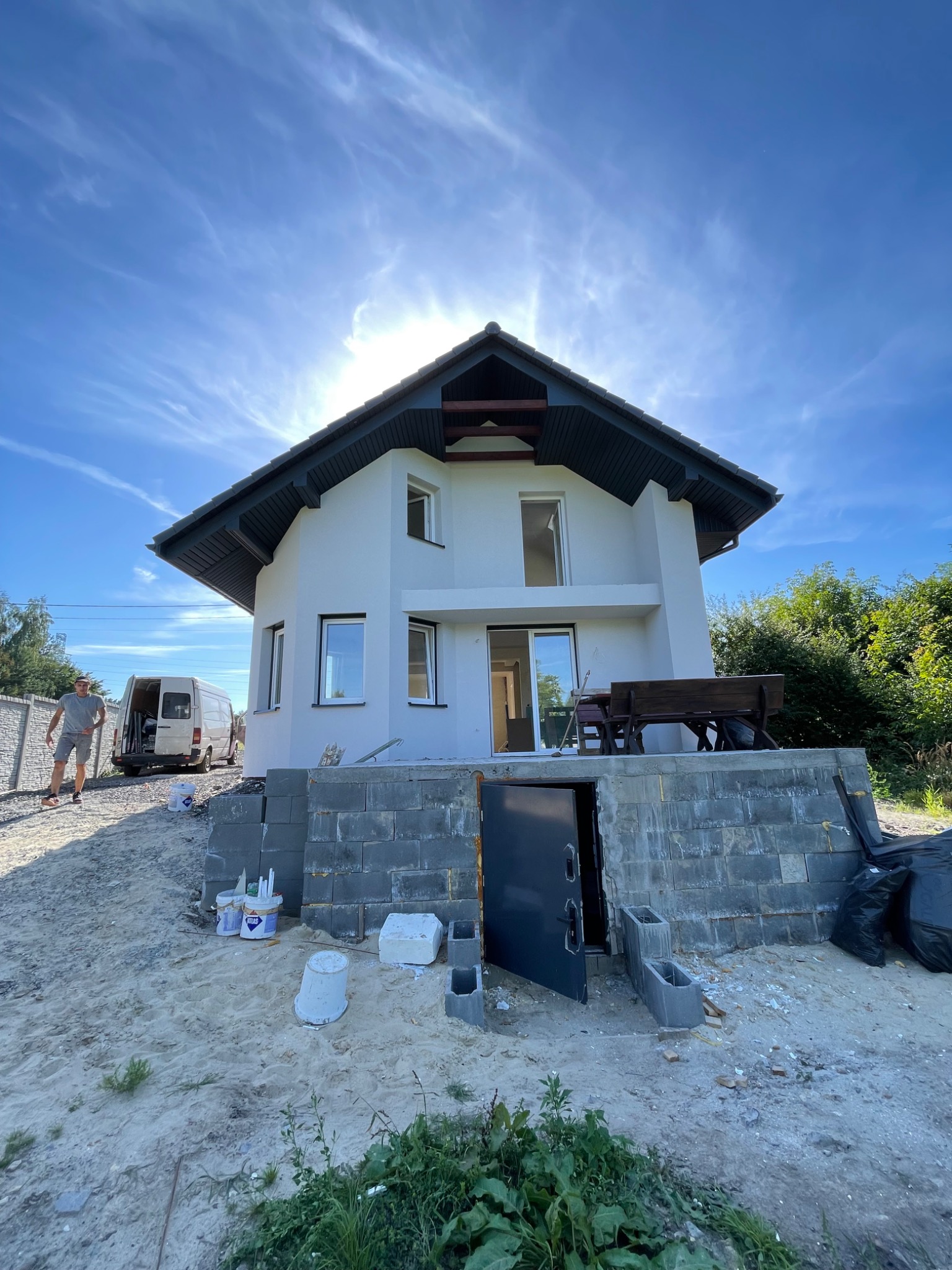 Dom jednorodzinny po tynkowaniu elewacji na etapie wykończeniowym, widoczne niedokończone fundamenty z bloczków betonowych, teren wokół w trakcie prac porządkowych, na tle błękitne niebo...