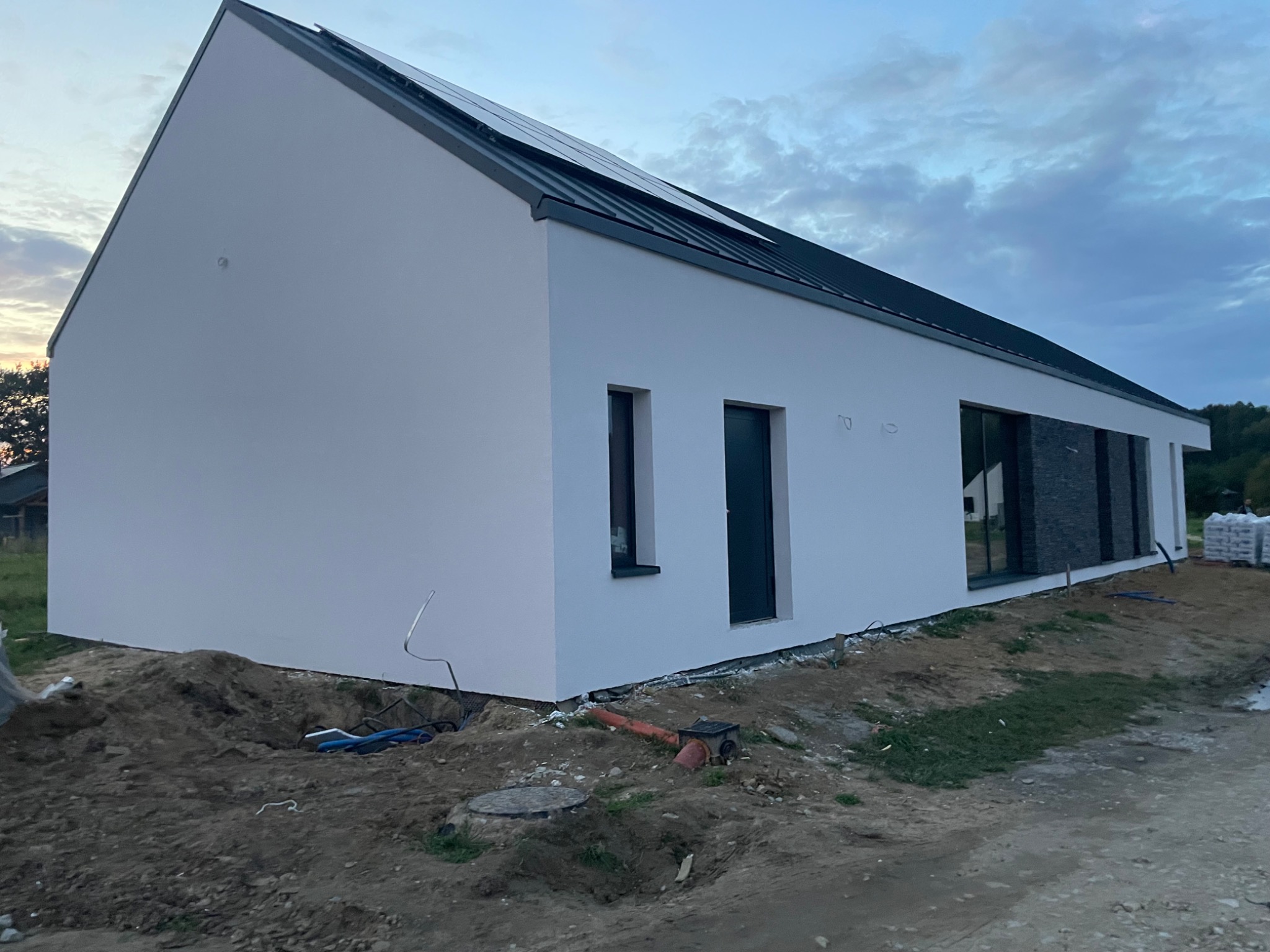 Nowoczesny dom jednorodzinny z białym tynkiem elewacyjnym i panelami słonecznymi na dachu, widok z zewnątrz, teren wokół w trakcie prac ziemnych.