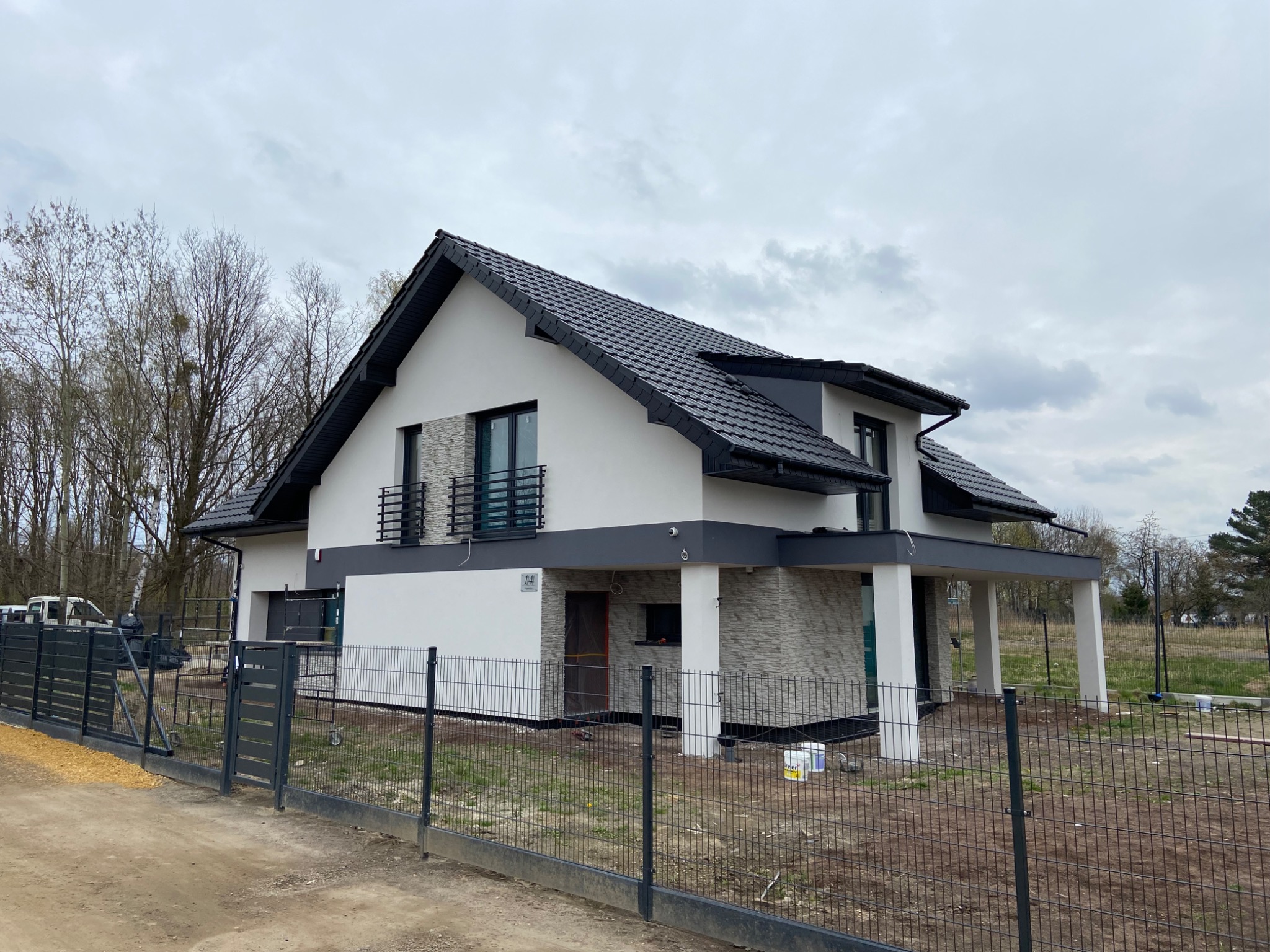 Nowoczesny dom jednorodzinny z białymi ścianami, szarą podmurówką i grafitowym dachem, otoczony metalowym ogrodzeniem panelowym na działce w trakcie zagospodarowywania.