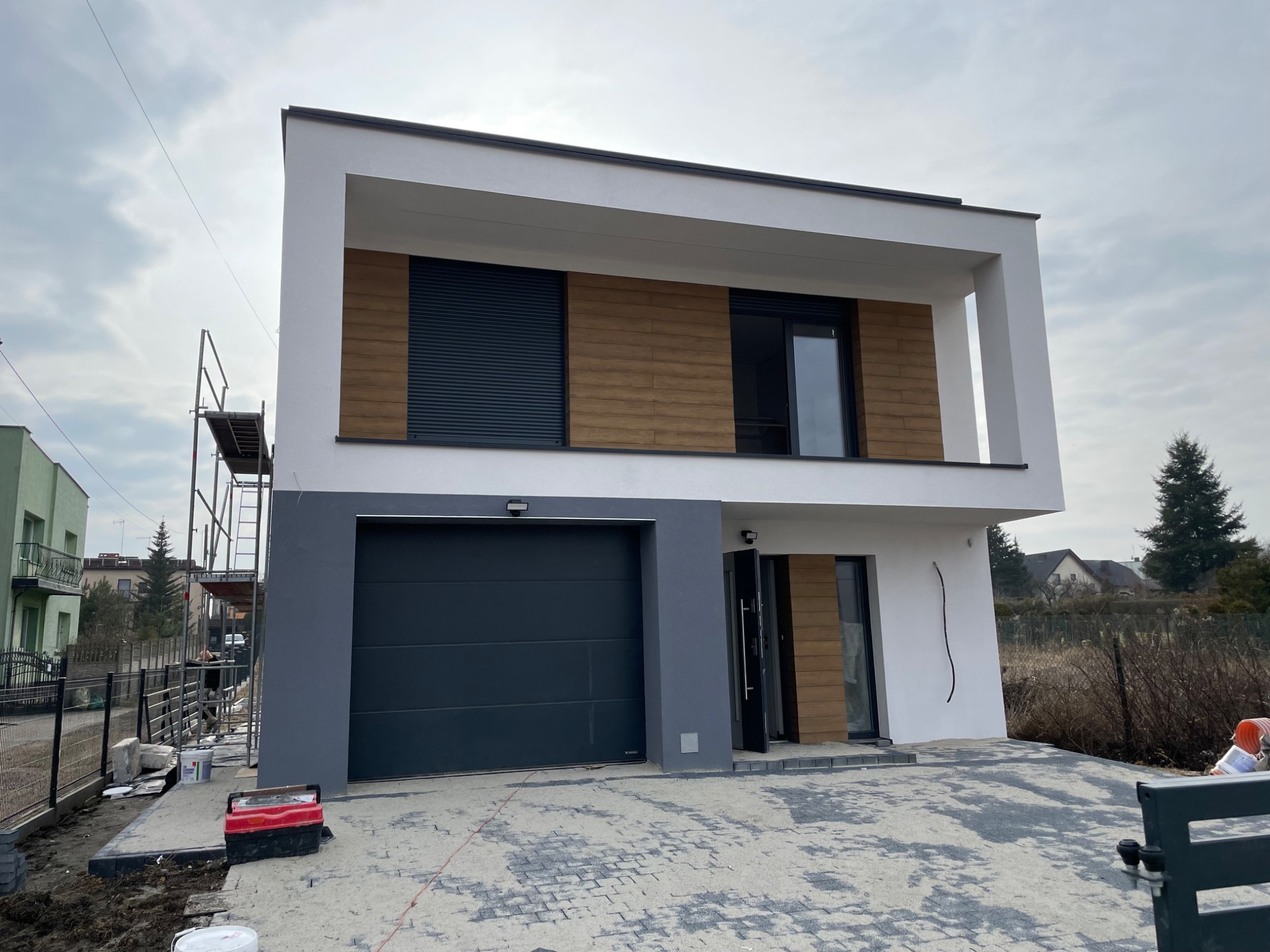 Nowoczesny dom dwukondygnacyjny z szarą bramą garażową i elementami drewnopodobnymi na elewacji, częściowo pomalowany, z widocznymi rusztowaniami i narzędziami na posesji.