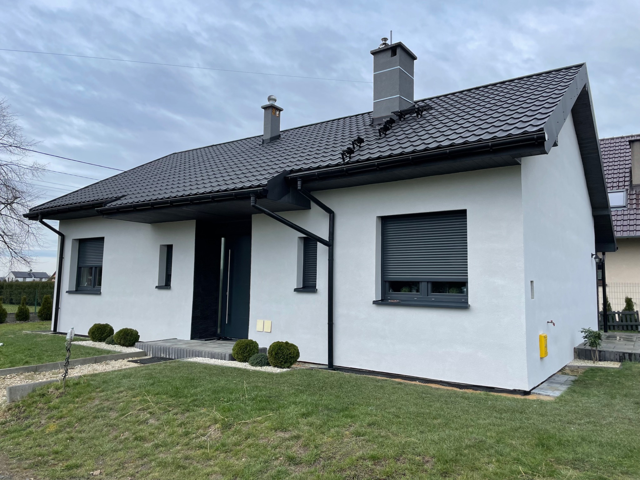 Nowoczesny dom jednorodzinny z białą elewacją, szarą dachówką i grafitowymi oknami z roletami zewnętrznymi, widok z trawnika z ozdobnymi krzewami.