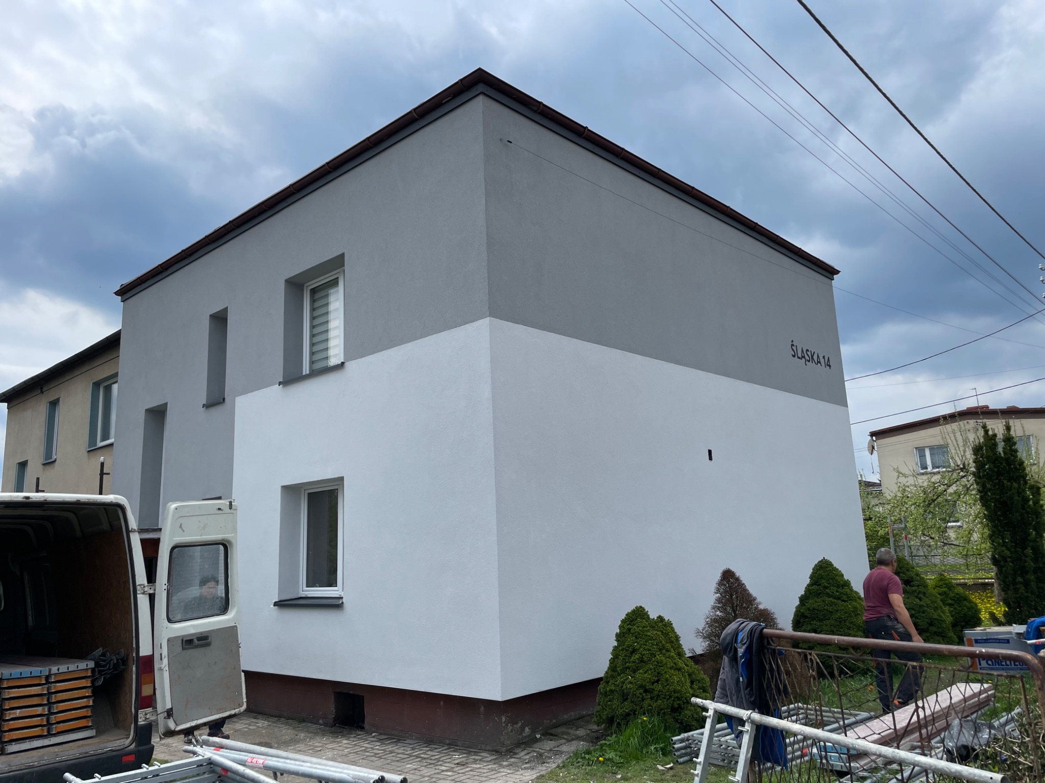 Dwukondygnacyjny budynek po częściowym odnowieniu elewacji, pomalowany na szaro i biało, z zaparkowanym vanem i rozłożonym rusztowaniem, widok z ulicy Śląska 14.