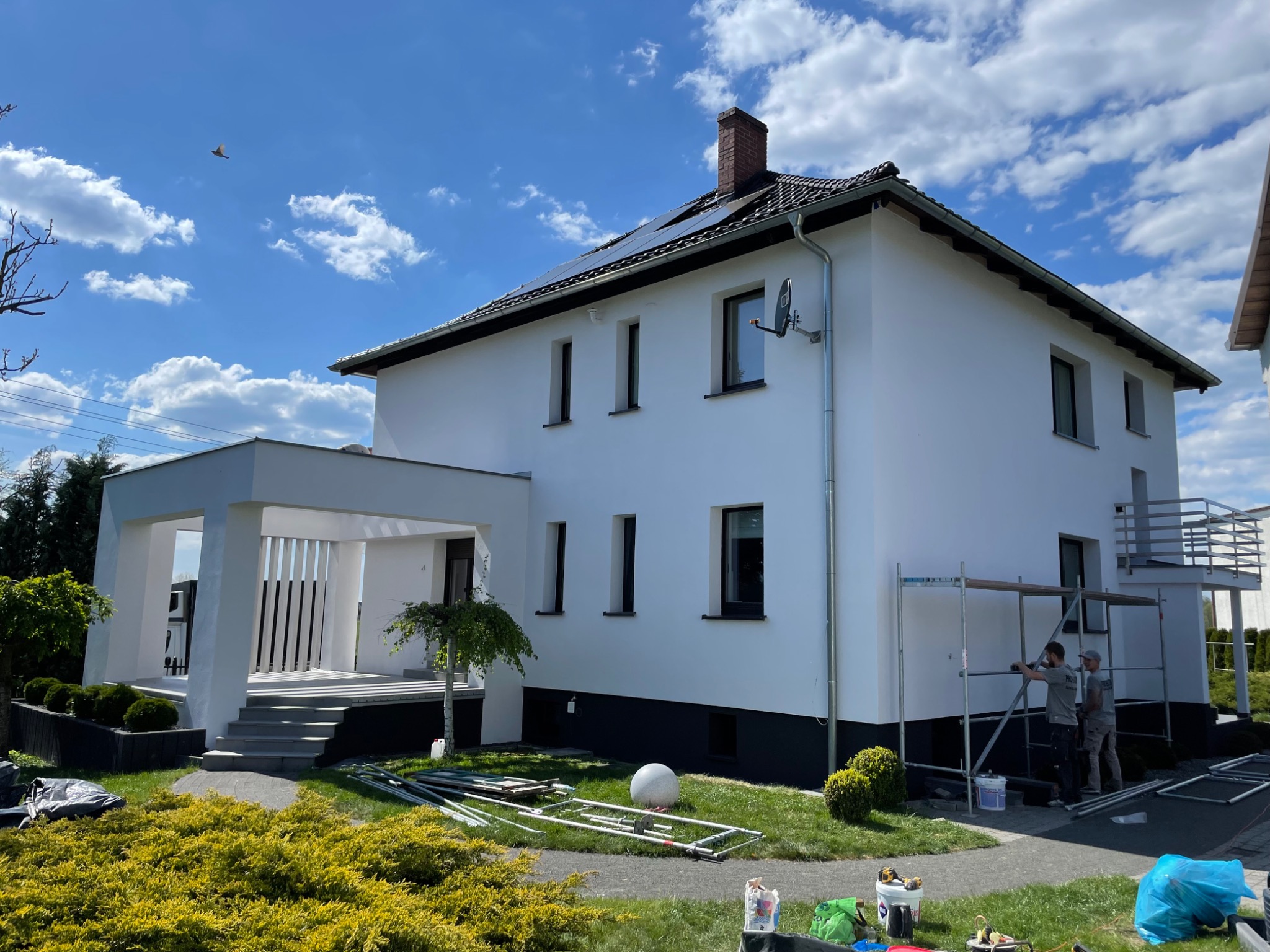 Dwóch pracowników na rusztowaniu przy białej elewacji domu w Chwostku, widoczne narzędzia i materiały budowlane na trawniku, błękitne niebo z obłokami.