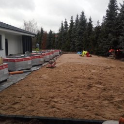 Kos-Tek - Przygotowanie terenu pod kostkę w Poznaniu: wyrównywanie piasku na dużej powierzchni z użyciem zagęszczarki i narzędzi ręcznych, widoczne ułożone obrzeża i palety z kostką.