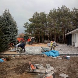 Kos-Tek - Ekipa brukarzy w trakcie prac: dwóch mężczyzn układa szarą kostkę na podsypce, w tle widoczny traktor, taczka, budynek mieszkalny i drzewa iglaste, na pierwszym planie dwie piły do cięcia kostki...