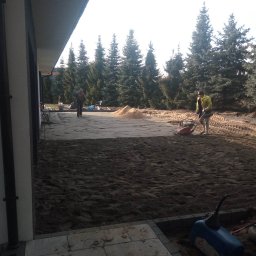 Kos-Tek - Prace ziemne w ogrodzie: ubijanie piasku zagęszczarką w przygotowaniu podłoża pod przyszłą nawierzchnię, widoczny piasek, drzewa w tle oraz fragment ułożonej kostki.