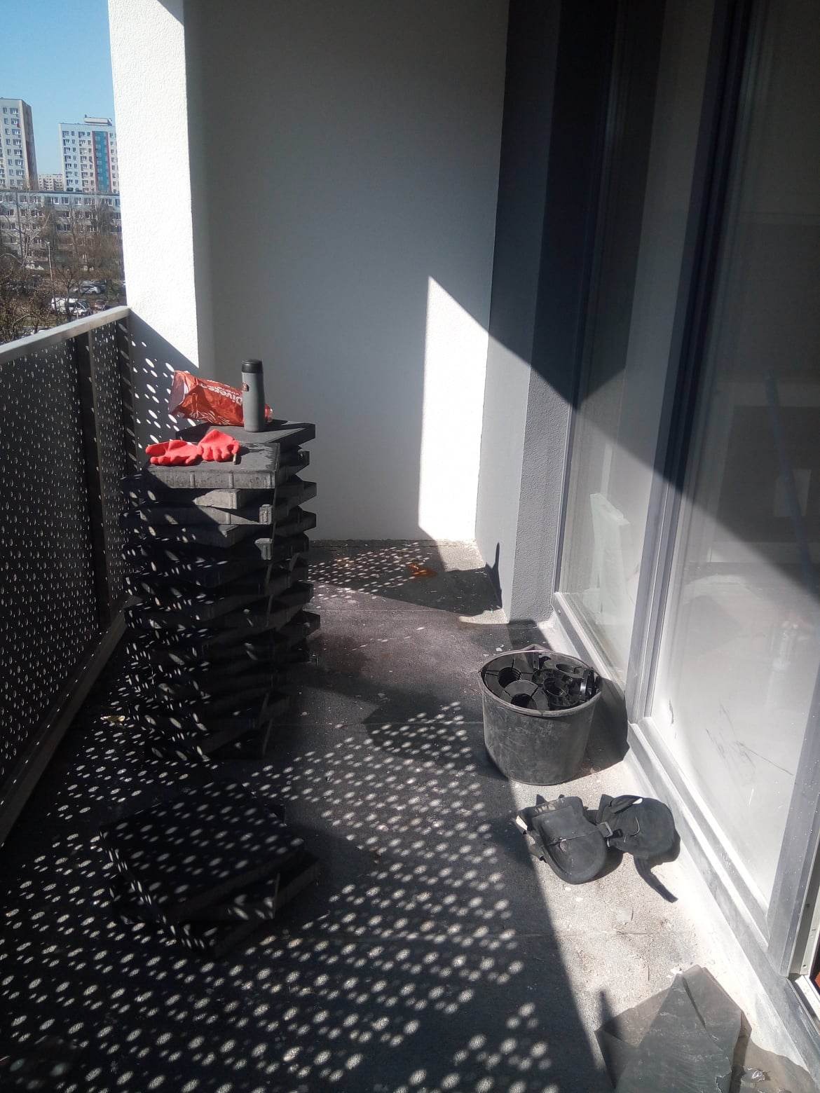 Stos szarych kostek brukowych na balkonie w bloku, obok wiadro z elementami i robocze buty, na wierzchu kostek czerwone rękawice i termos.
