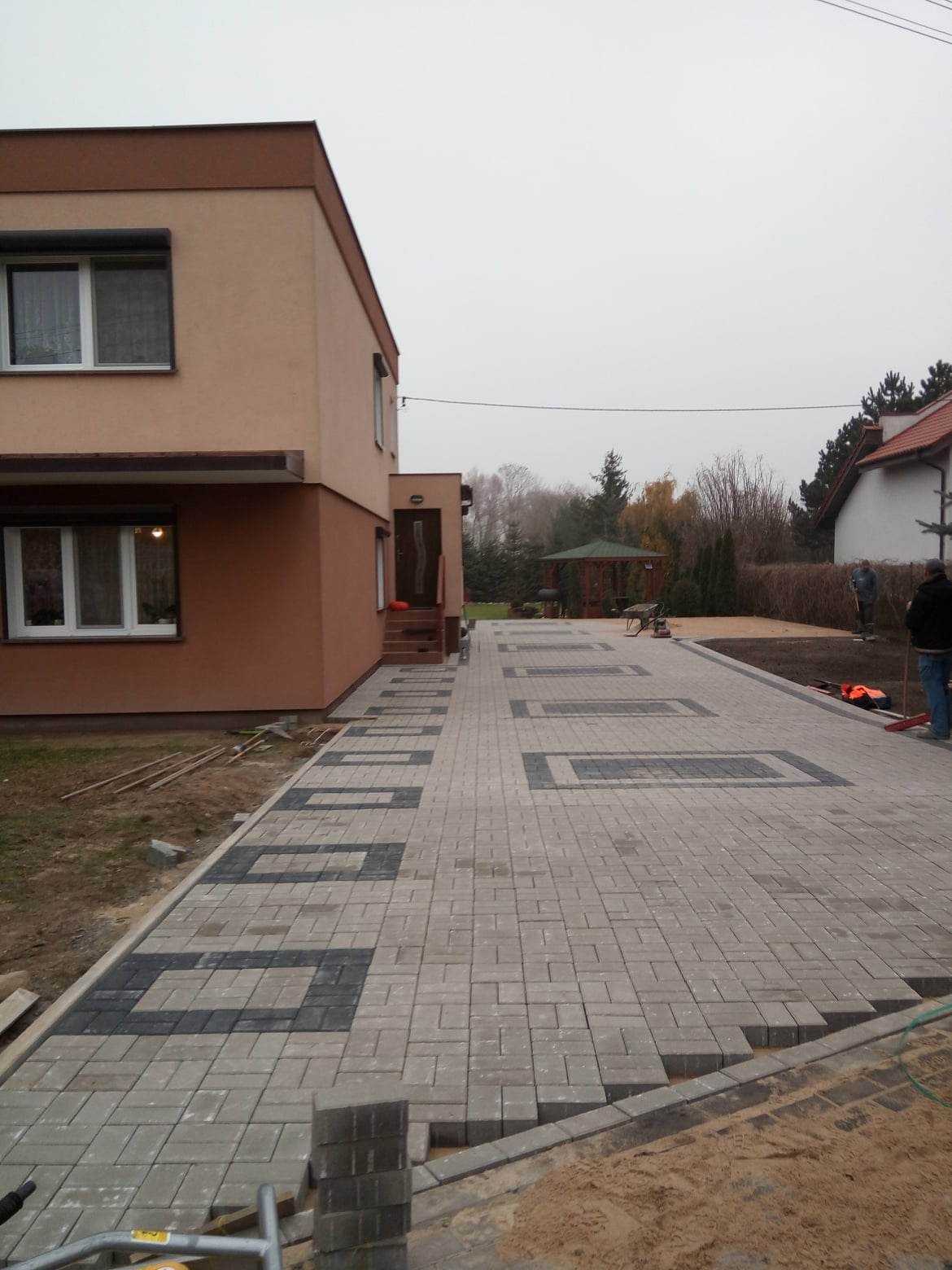 Nowo ułożony podjazd z kostki brukowej w odcieniach szarości z ciemnymi wstawkami, prowadzący do domu z brązową elewacją; widoczni pracownicy i narzędzia brukarskie w tle.