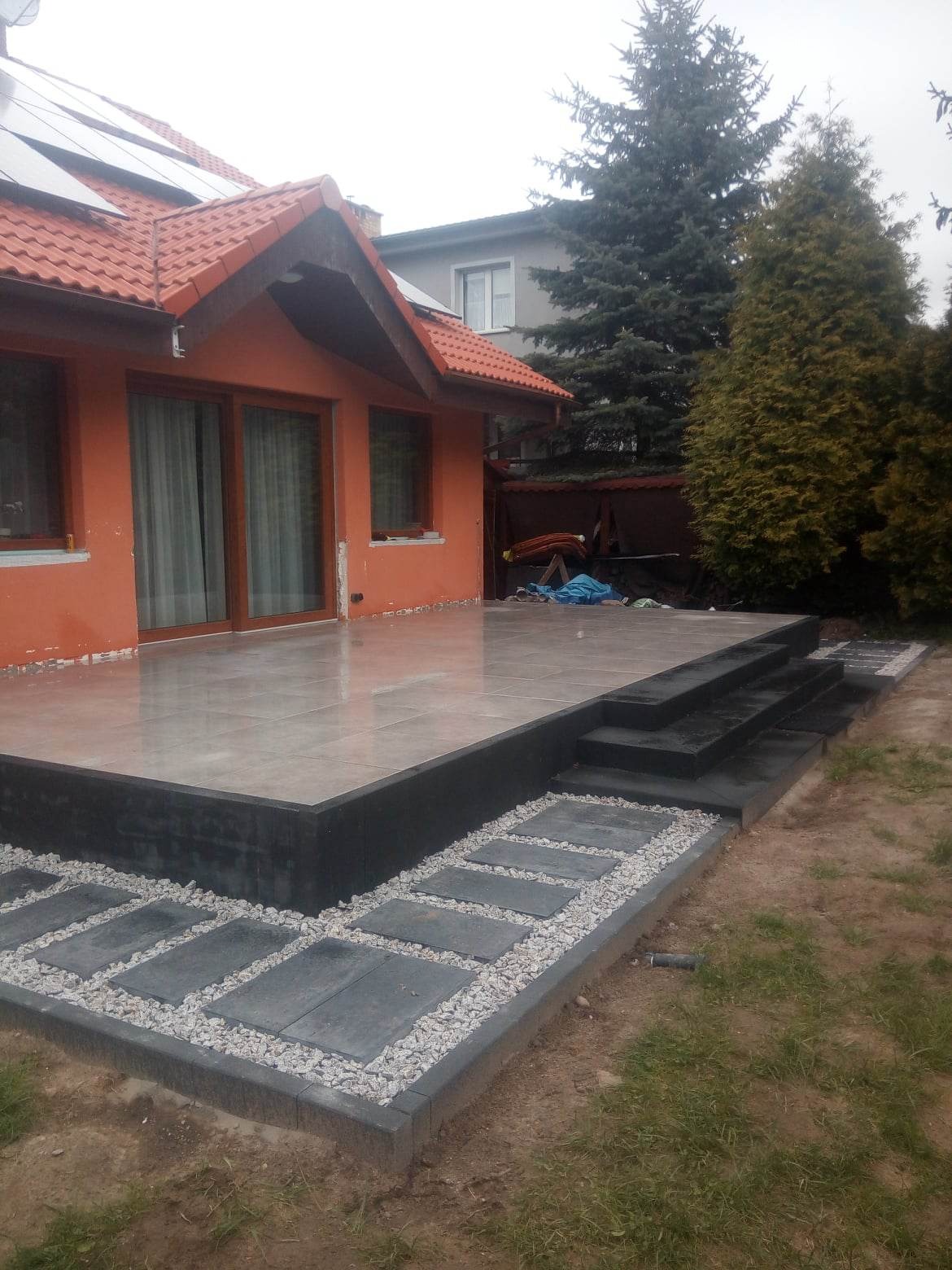 Wykończony taras z szarych płytek ceramicznych, otoczony czarną obudową i schodami, z dekoracyjną ścieżką z kamieni i płyt w ogrodzie.