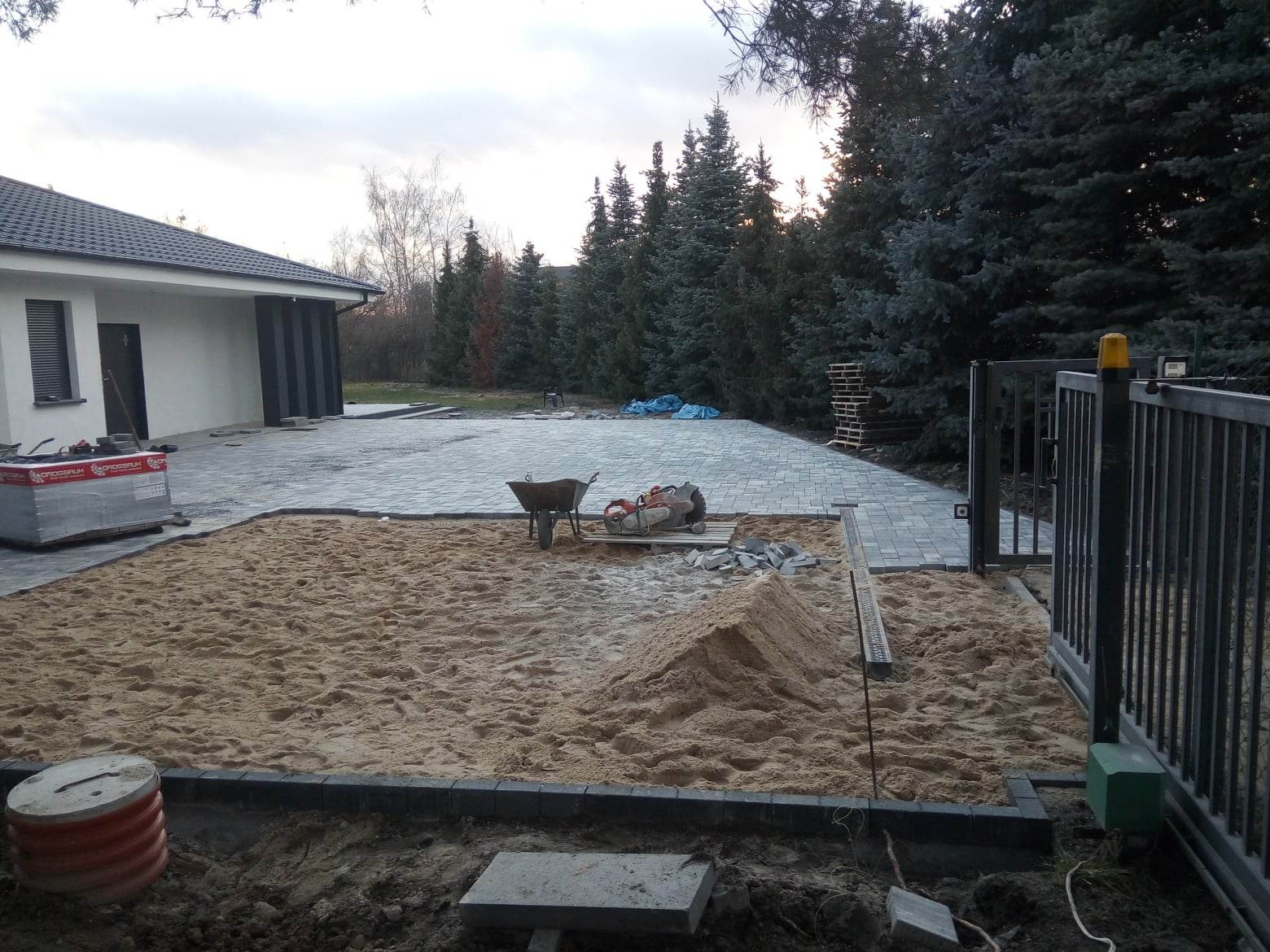 Teren przed domem w trakcie przygotowania podłoża pod nawierzchnię z kostki brukowej, widoczny piasek, krawężniki, częściowo ułożona kostka, taczka, przecinarka do kostki i paleta z kostką.
