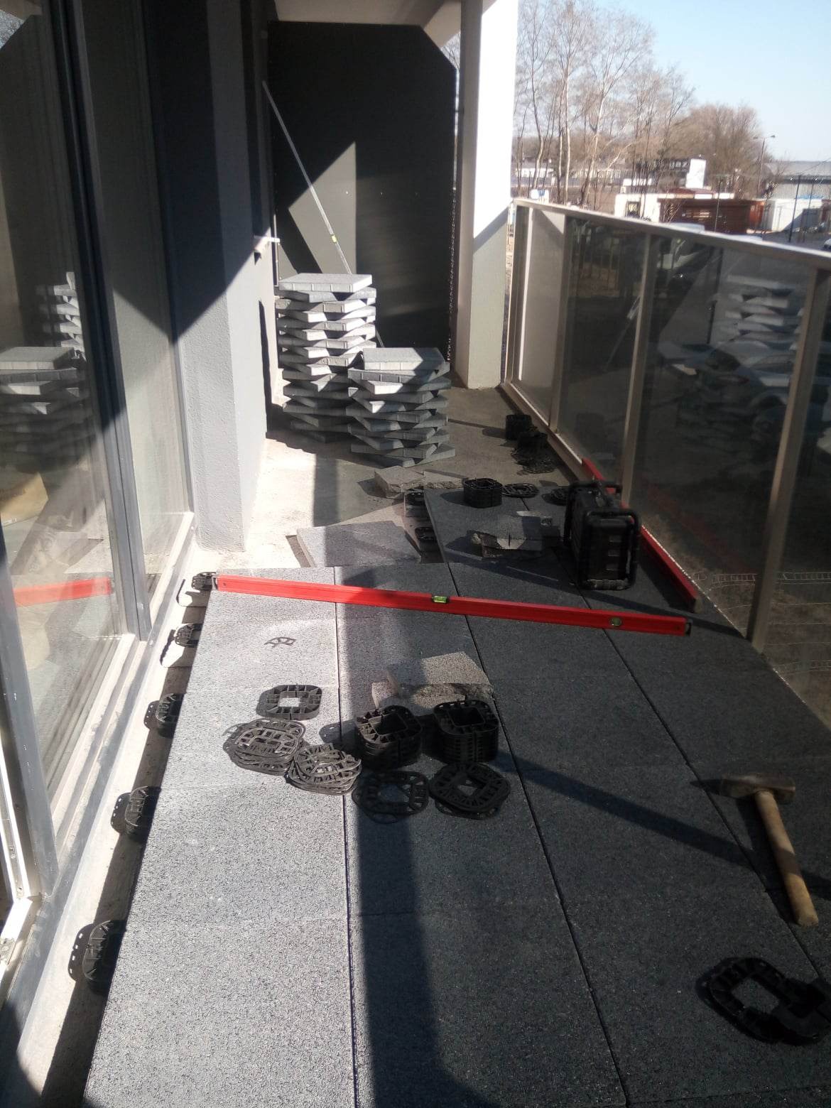 Montaż podłogi na balkonie: płyty na dystansach, poziomica i młotek w kadrze, w tle stos kostki.