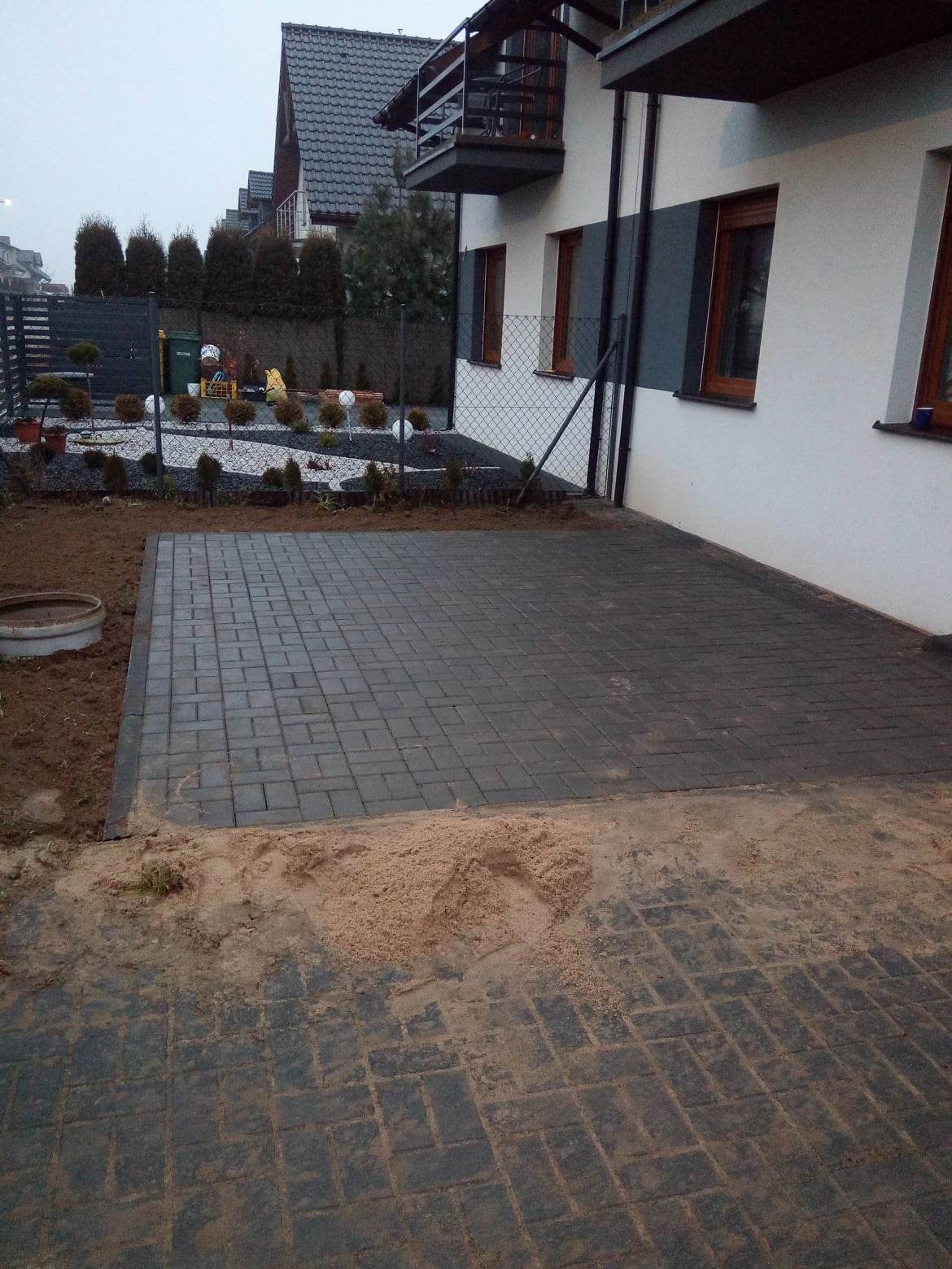 Częściowo ułożona, grafitowa kostka brukowa przed nowoczesnym domem jednorodzinnym; widoczna warstwa piasku pod przyszłą nawierzchnią, ogrodzenie siatkowe i zagospodarowany ogród w tle.