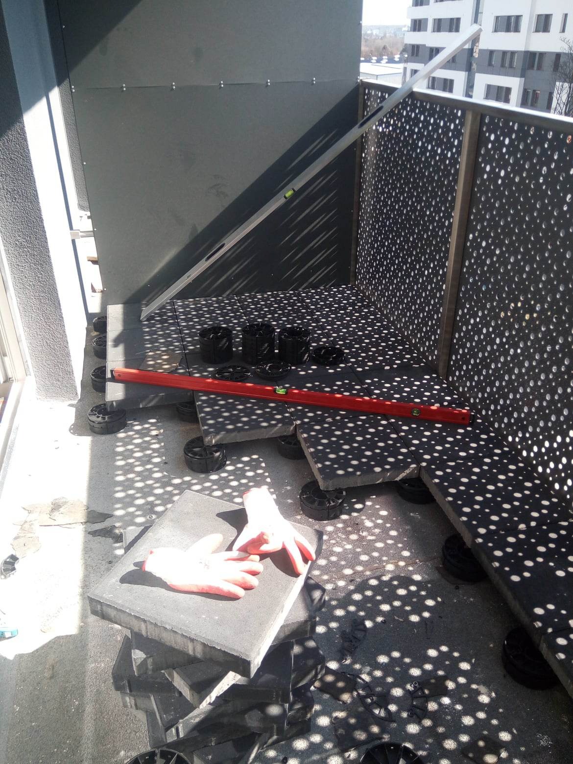 Montaż podłogi na balkonie z użyciem regulowanych wsporników, poziomicy i roboczych rękawic; widoczne cienie rzucane przez ażurowe panele balkonowe.