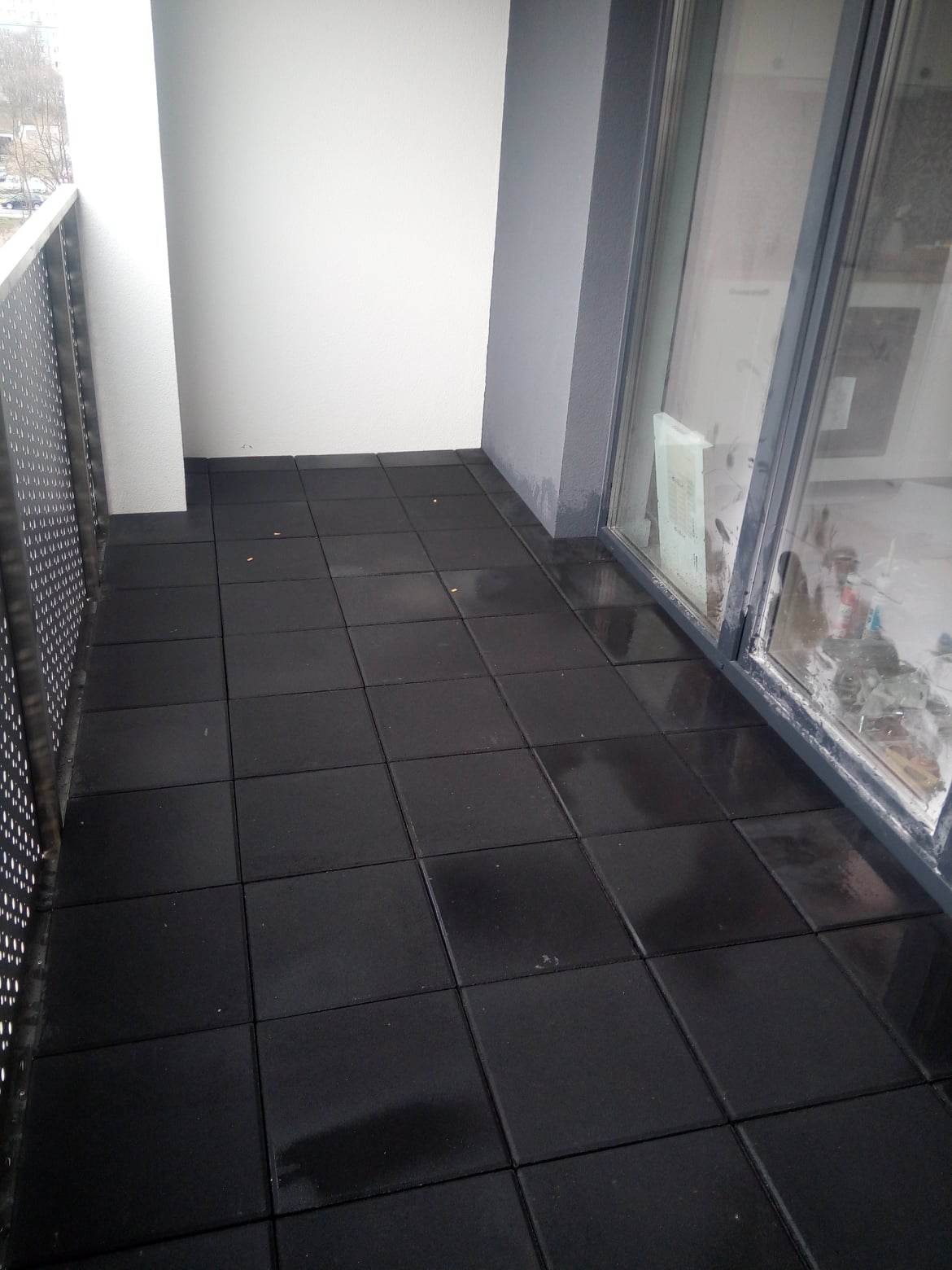 Czarne, kwadratowe płyty ułożone na balkonie, widoczne metalowe barierki i przesuwne drzwi balkonowe.