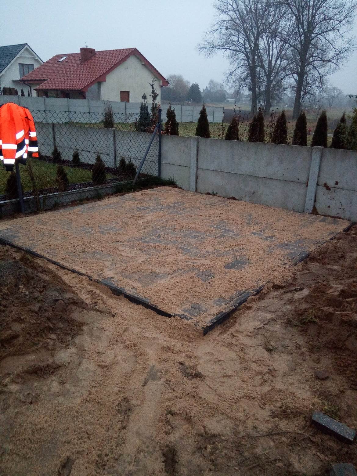 Przygotowany teren pod kostkę brukową, podsypany piaskiem, z widoczną pomarańczową kurtką odblaskową na ogrodzeniu w tle.