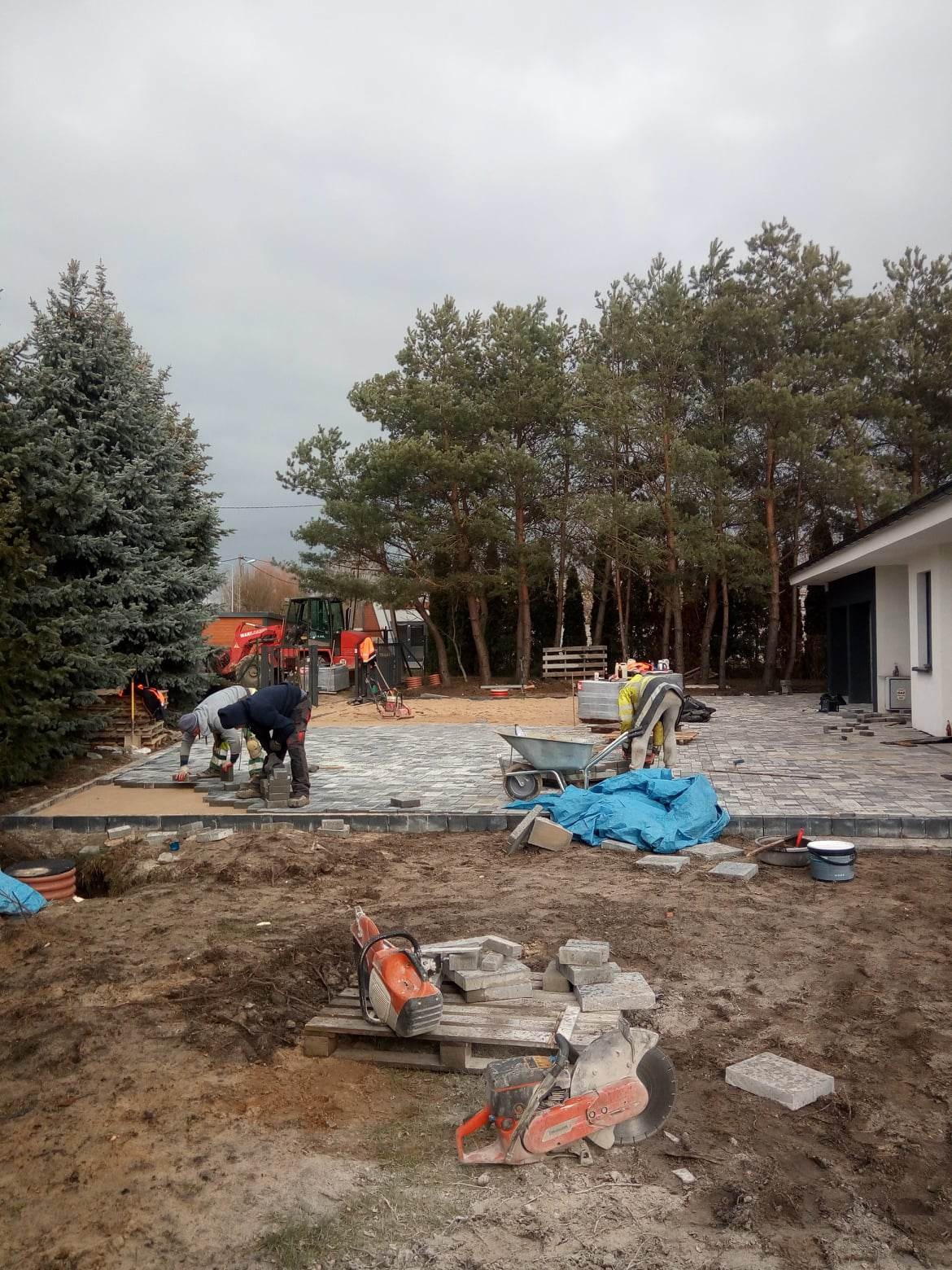 Ekipa brukarzy w trakcie prac: dwóch mężczyzn układa szarą kostkę na podsypce, w tle widoczny traktor, taczka, budynek mieszkalny i drzewa iglaste, na pierwszym planie dwie piły do cięcia kostki...