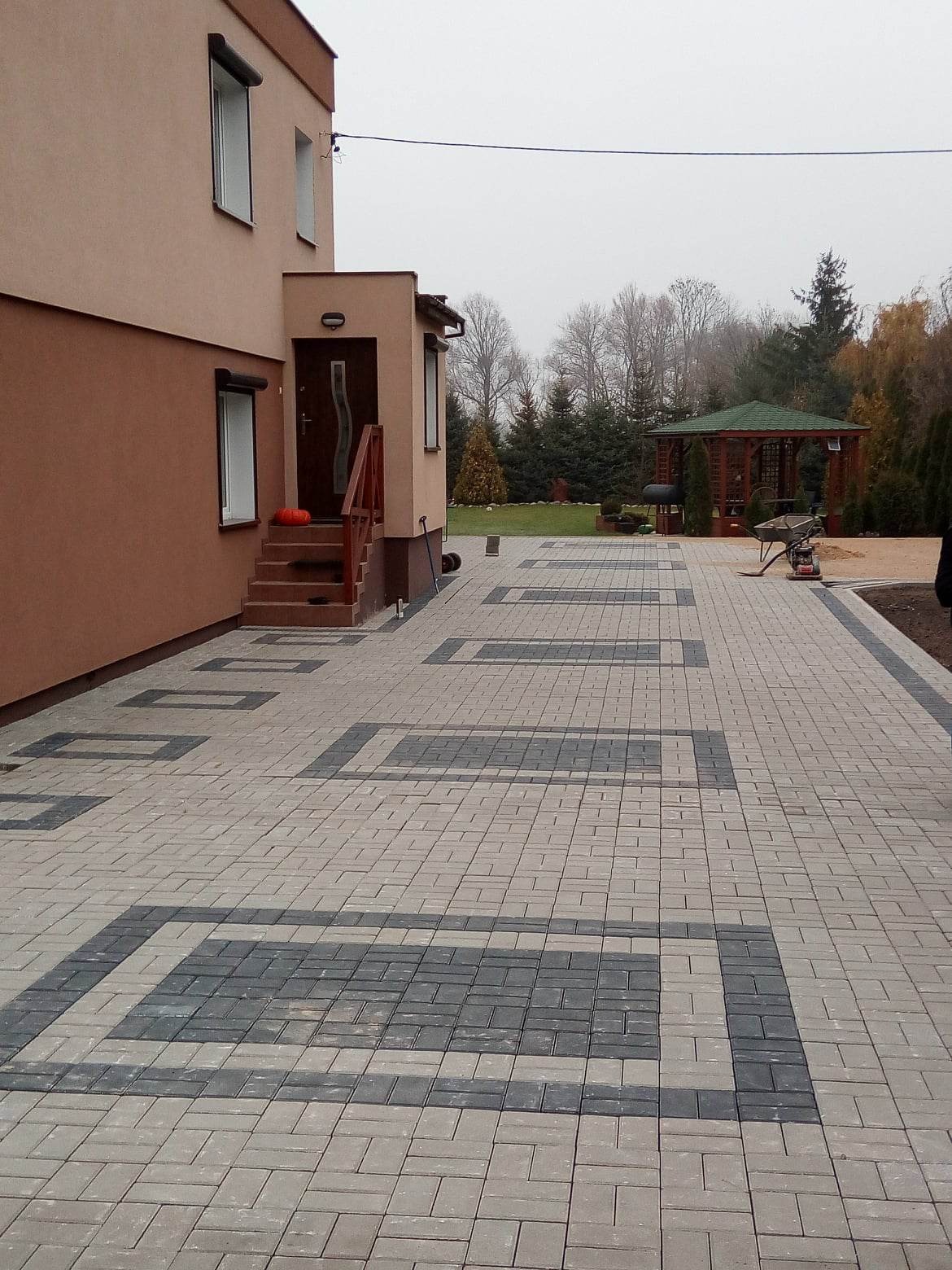 Ułożona kostka brukowa w geometryczny wzór przed domem z drewnianą altaną w tle, widoczne schody wejściowe z dekoracyjną dynią.