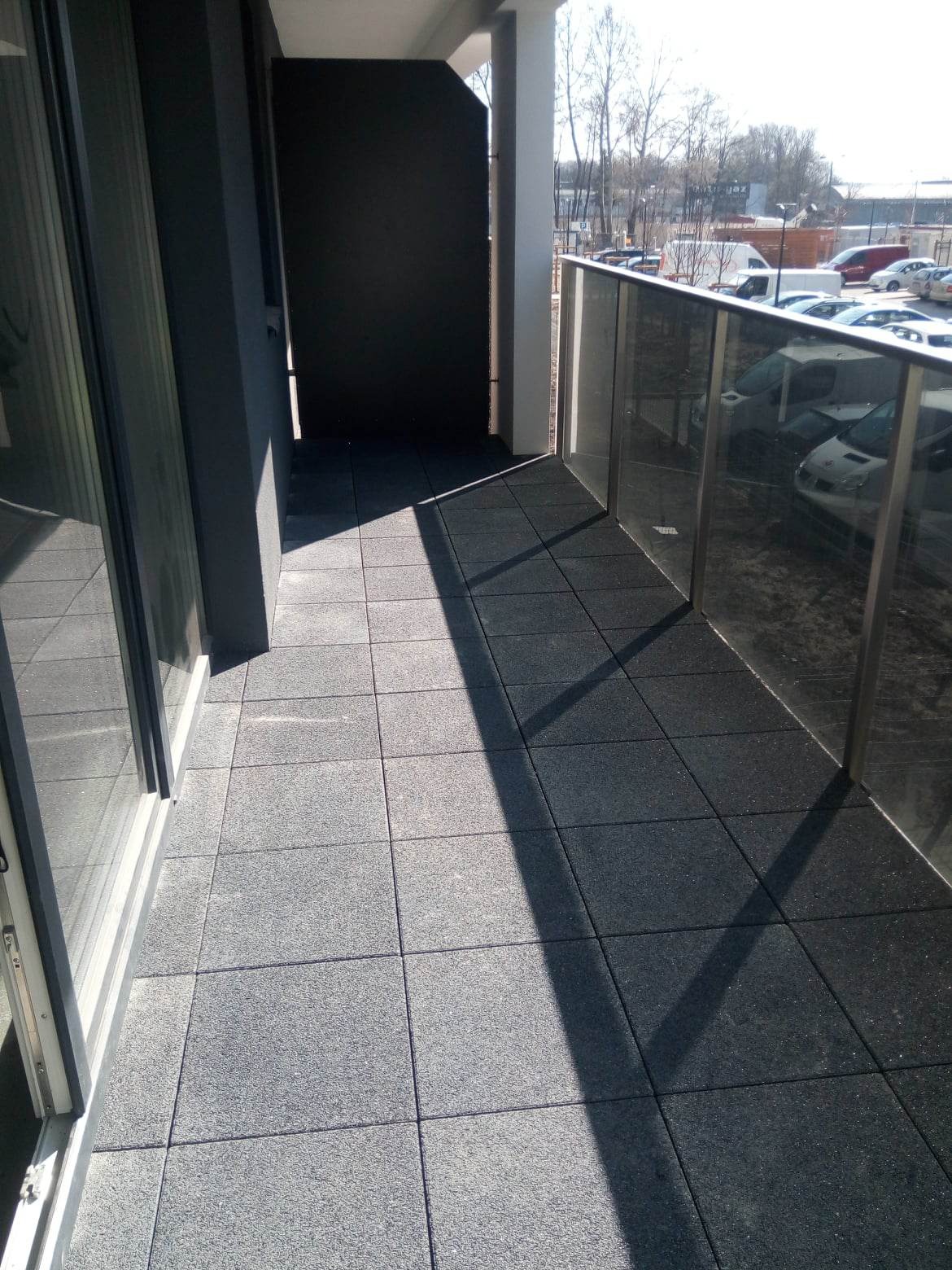 Wykończony balkon z szarą, gumową kostką brukową, szklaną balustradą i widokiem na parking.