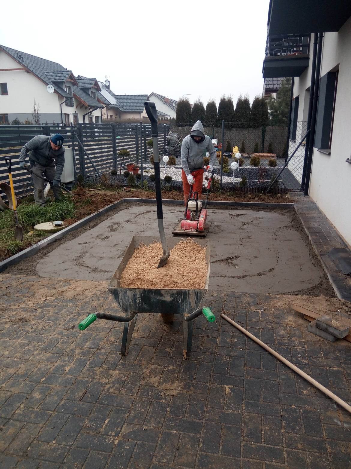 Przygotowanie podłoża pod kostkę brukową: ubijanie piasku zagęszczarką, widoczna taczka z piaskiem, pracownicy w trakcie robót ziemnych na posesji.