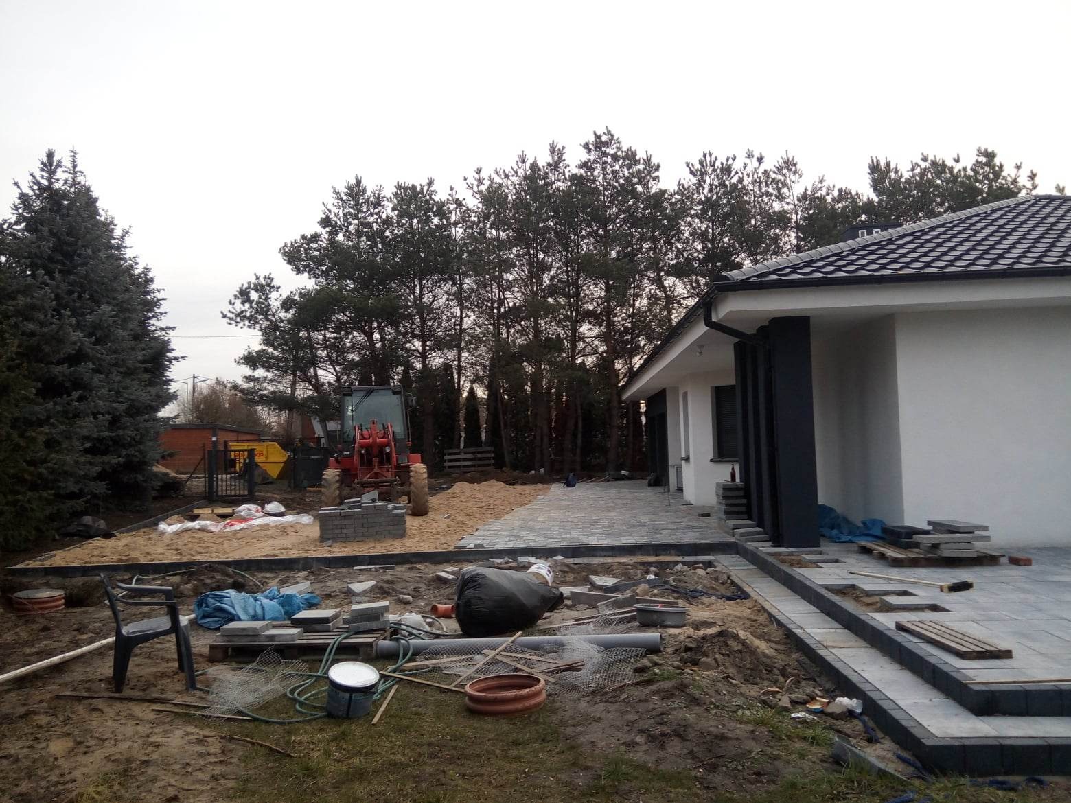 Układanie szarej kostki brukowej na posesji z nowoczesnym domem; widoczny czerwony traktor, piasek, narzędzia i częściowo ułożona nawierzchnia.