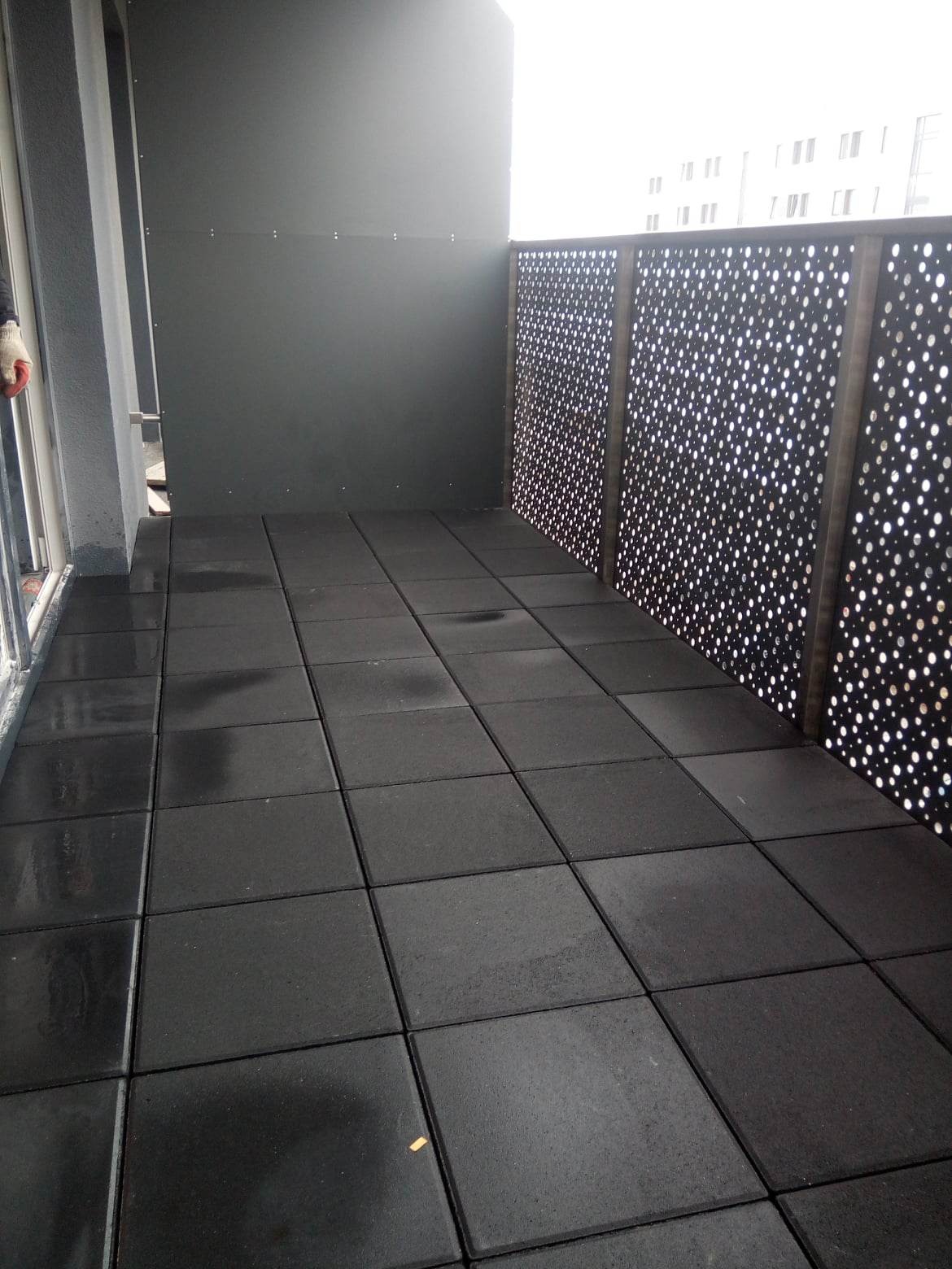 Ciemnoszare, gumowe płyty na balkonie z ażurową balustradą i szarą ścianą w tle.