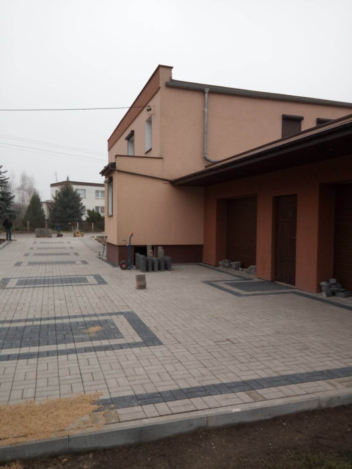 Ułożona kostka brukowa w geometryczny wzór przed budynkiem mieszkalnym z garażem, widoczne palety z kostką i wózek transportowy.