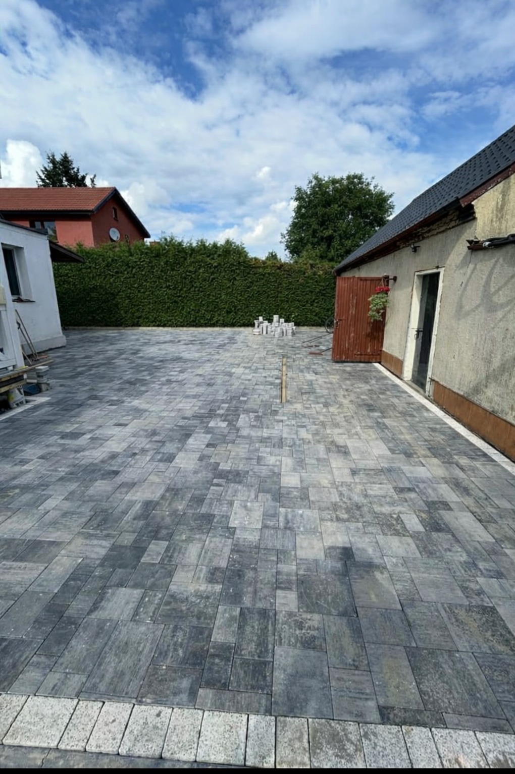 Ułożona kostka brukowa w Śmiłowie, wzór szaro-czarny, obrzeża z jasnej kostki. Widoczne budynki w tle i zielony żywopłot. Prace brukarskie.