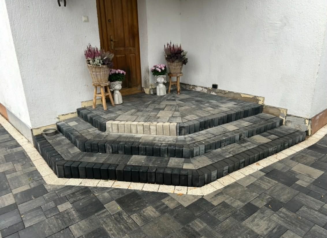 Schody wejściowe z ciemnej kostki brukowej, wykończone jaśniejszą obwódką. Dekoracyjne donice z kwiatami przy drzwiach. Estetyczne i funkcjonalne rozwiązanie.