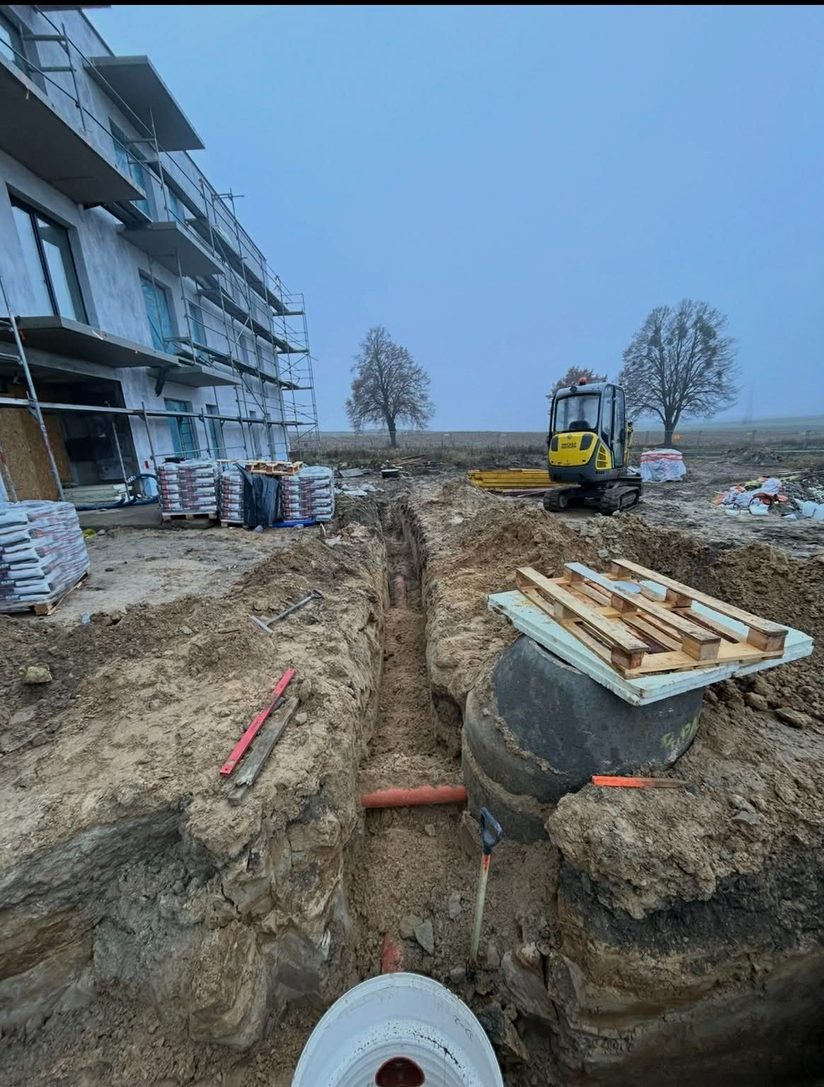 Wykopany rów z widocznymi rurami, mini koparka w tle, budynek w stanie surowym i palety z materiałami budowlanymi. Prace ziemne na budowie.