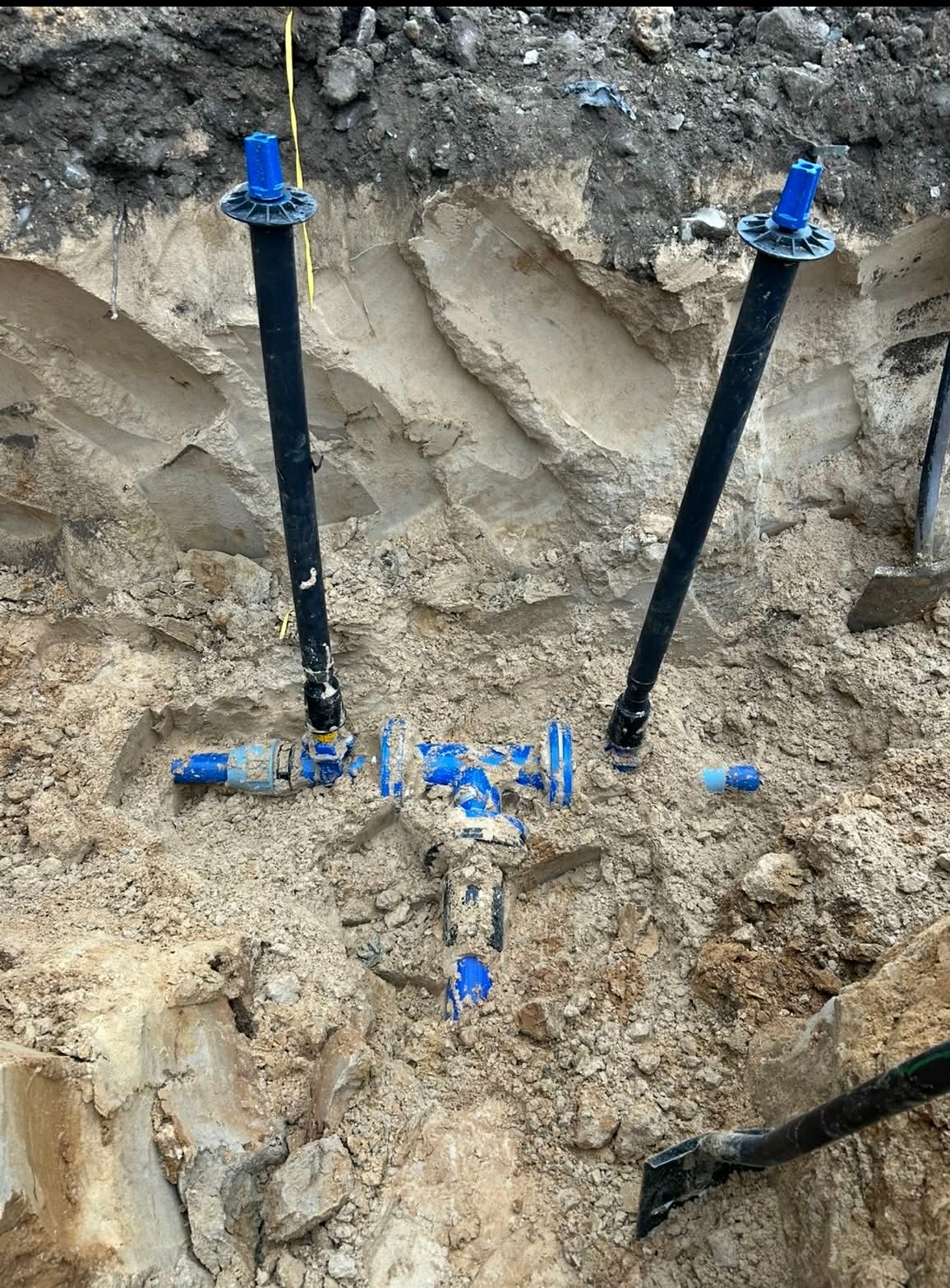 Wykop z widocznymi elementami instalacji wodociągowej: rury pionowe z niebieskimi zakończeniami, niebieskie złączki i zawory. Widoczne ślady ziemi i narzędzia.
