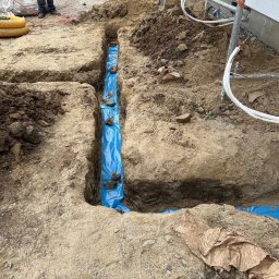 Elektro-Automatic Marcin Michalak - Wykop w ziemi wyłożony niebieską folią, prawdopodobnie pod instalację, z widocznymi przewodami elektrycznymi przy budynku w tle.