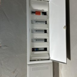 Elektro-Automatic Marcin Michalak - Otwarta, biała skrzynka rozdzielcza z bezpiecznikami i wyłącznikami zamontowana na surowej, szarej ścianie. Widoczne numery obwodów. Dolna część skrzynki zamknięta, z perforowaną obudową.