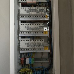 Elektro-Automatic Marcin Michalak - Otwarta skrzynka bezpiecznikowa z widocznymi bezpiecznikami, przewodami i listwami zaciskowymi, zamontowana na szarej ścianie.