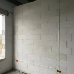 Surowa ściana z betonu komórkowego wewnątrz budynku w trakcie budowy, z widocznymi puszkami elektrycznymi i poprowadzonymi przewodami w białej rurce PCV.