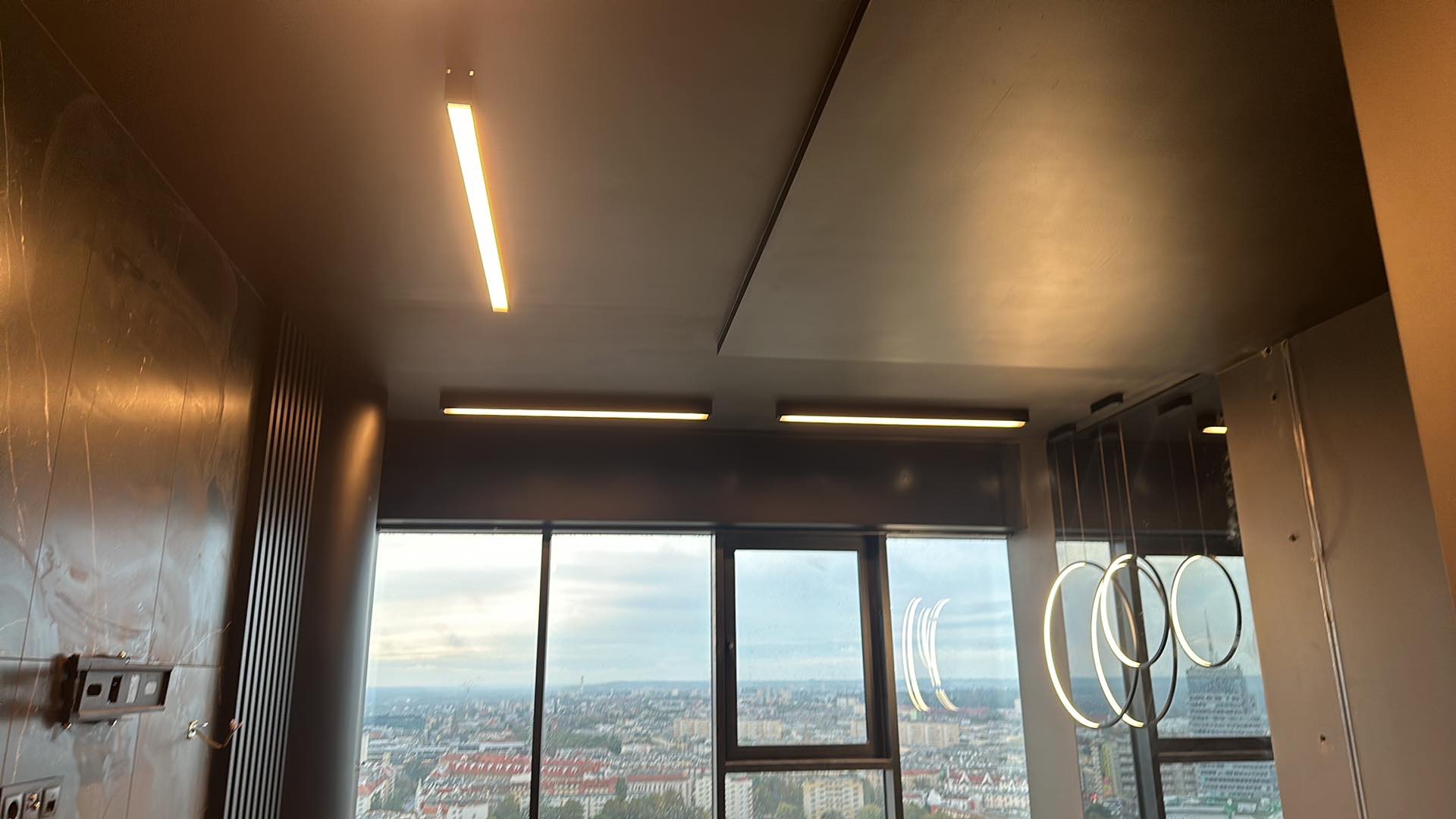 Nowoczesne oświetlenie sufitowe w apartamencie z widokiem na miasto, obejmujące liniowe lampy LED i wiszące okręgi świetlne.