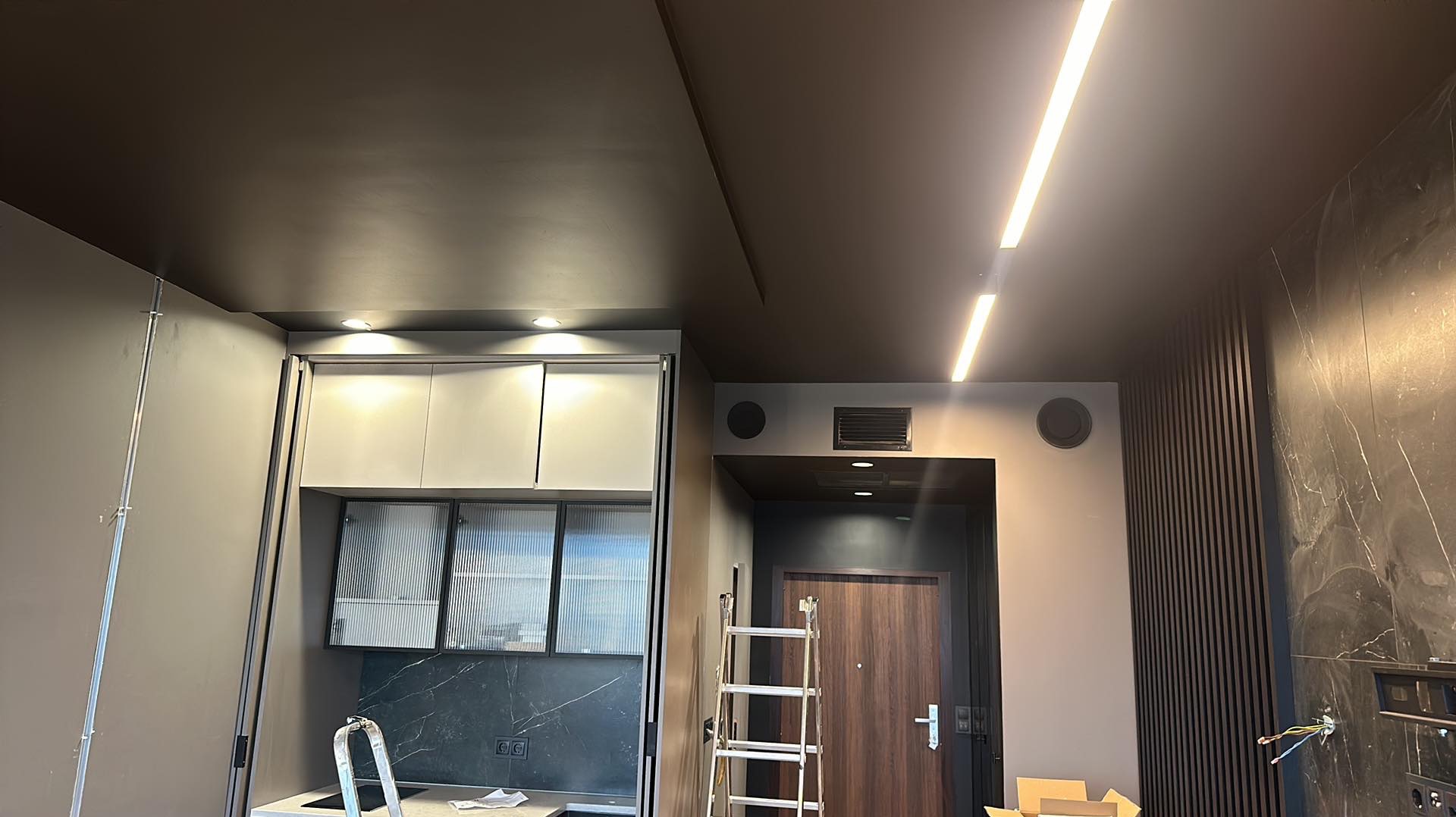 Nowoczesne wnętrze w trakcie wykańczania: widoczna drabina oparta o ścianę, zamontowane oświetlenie LED na suficie i halogeny, zabudowa kuchenna z ryflowanymi szybami, czarny sufit i elementy...