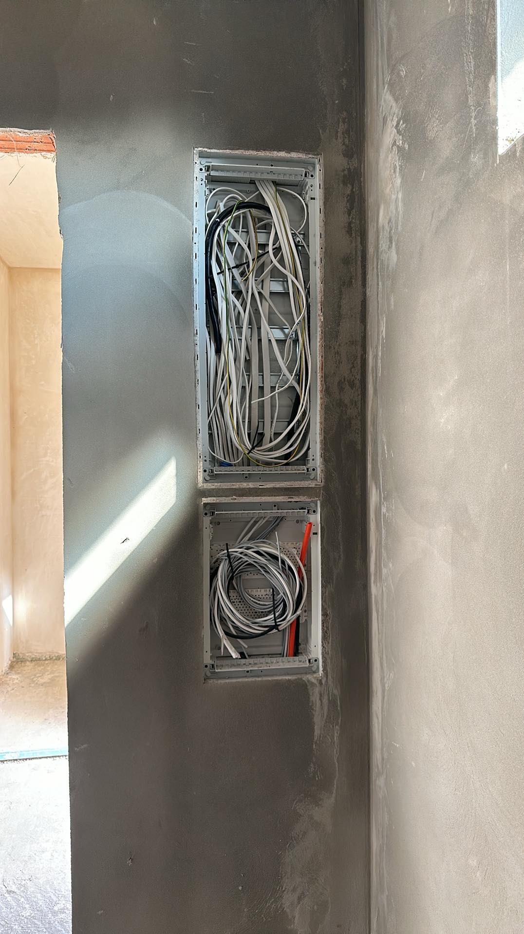 Dwa podtynkowe panele elektryczne z przewodami w ścianie w trakcie prac wykończeniowych, widoczne przewody białe, czarne i żółto-zielone.